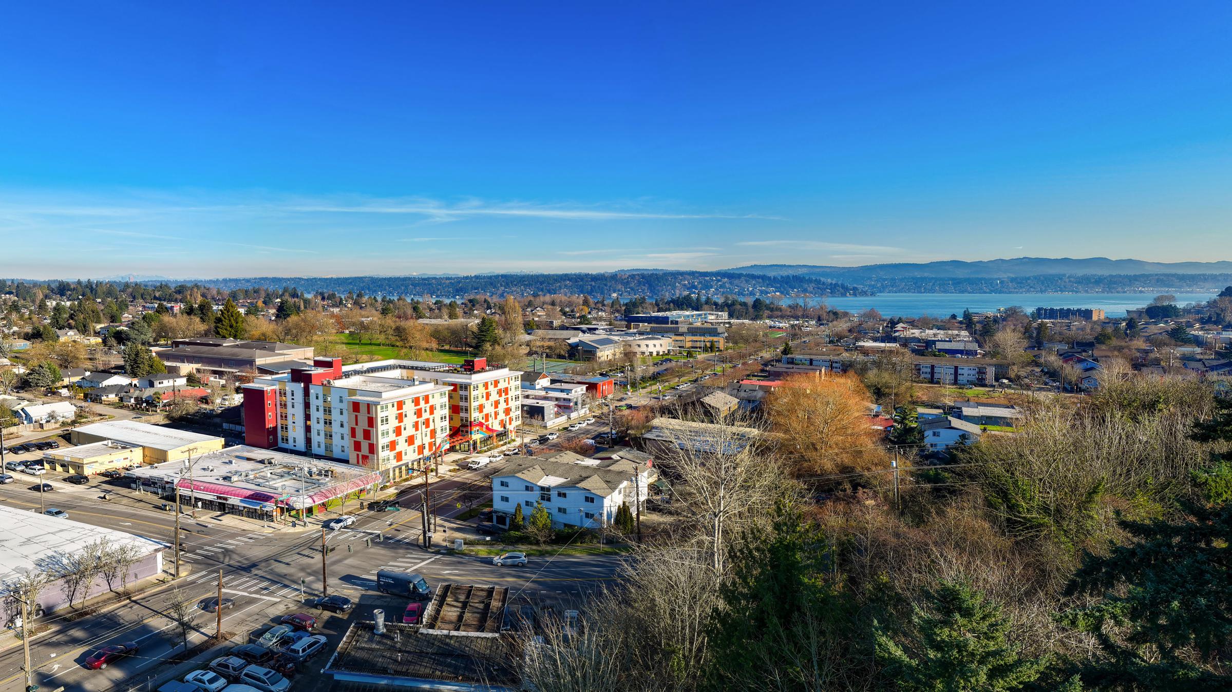A panoramic view of a suburban area featuring a mix of residential and commercial buildings, with a prominent red and white building in the foreground. The landscape includes tree-lined streets and distant hills, with a lake visible in the background under a clear blue sky.