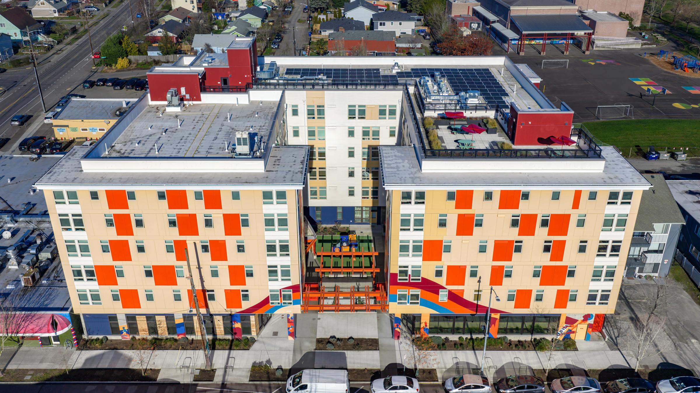 Aerial view of a modern, multi-story building with a colorful facade featuring orange and yellow panels. The building has a spacious entrance with outdoor seating and vibrant decor. Surrounding the structure are parked cars and nearby residential buildings, with a playground visible in the background.