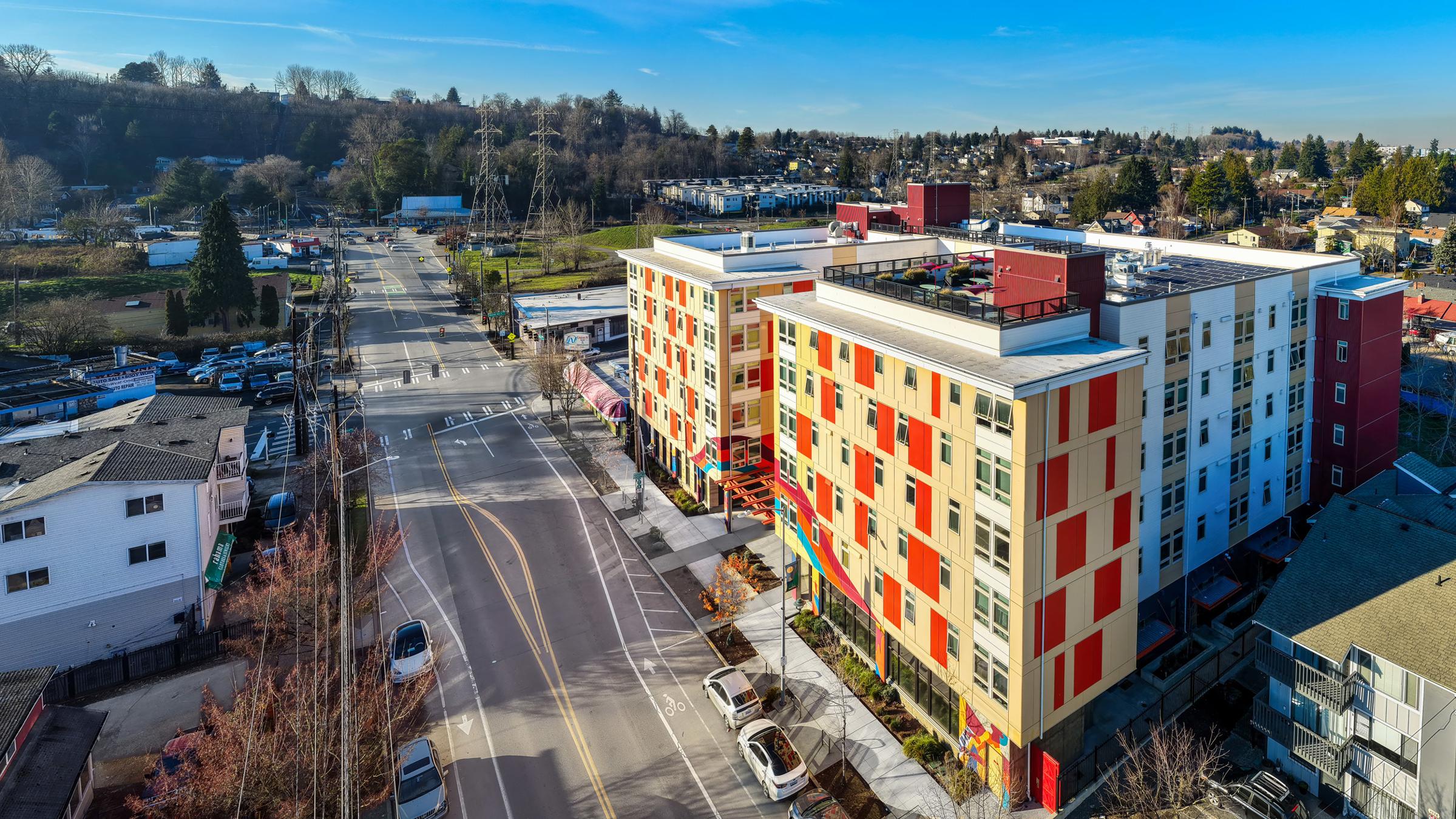 Aerial view of a modern multi-story building with a colorful facade, surrounded by residential and commercial areas. The scene features a wide street, parked cars, and trees in the background under a clear blue sky.