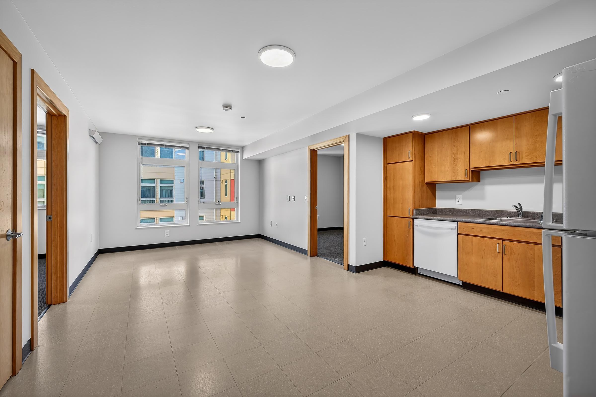 A modern, empty kitchen and living area featuring light-colored walls, laminate flooring, and wooden cabinetry. Large windows provide natural light, with one window overlooking an exterior view. The kitchen includes a white refrigerator and dishwasher, with a countertop and sink visible.