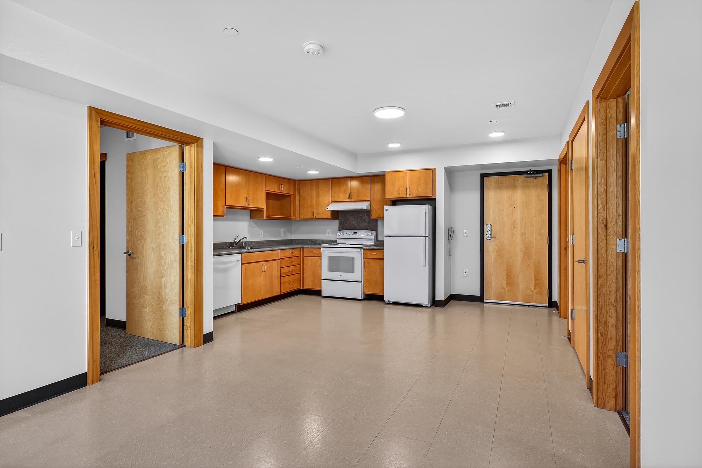Modern kitchen in an open layout showing wooden cabinets, a white refrigerator, and a stove. The room features light-colored walls and flooring, with two doors leading to other areas. Bright overhead lighting enhances the space, which is uncluttered and inviting.