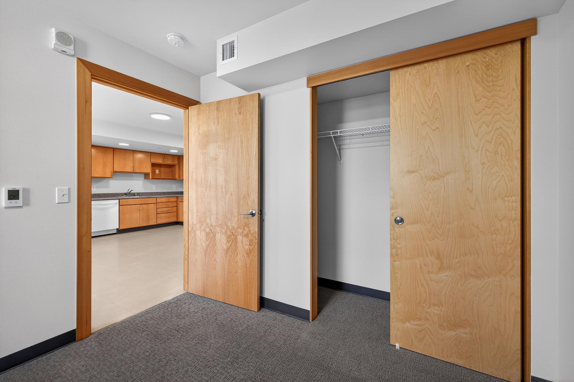 Interior view of a small room featuring two wooden doors, one leading to a closet with a shelf. The other door opens to a kitchen area with wooden cabinets and modern appliances visible in the background. The flooring is light-colored tile, and there is neutral wall paint.
