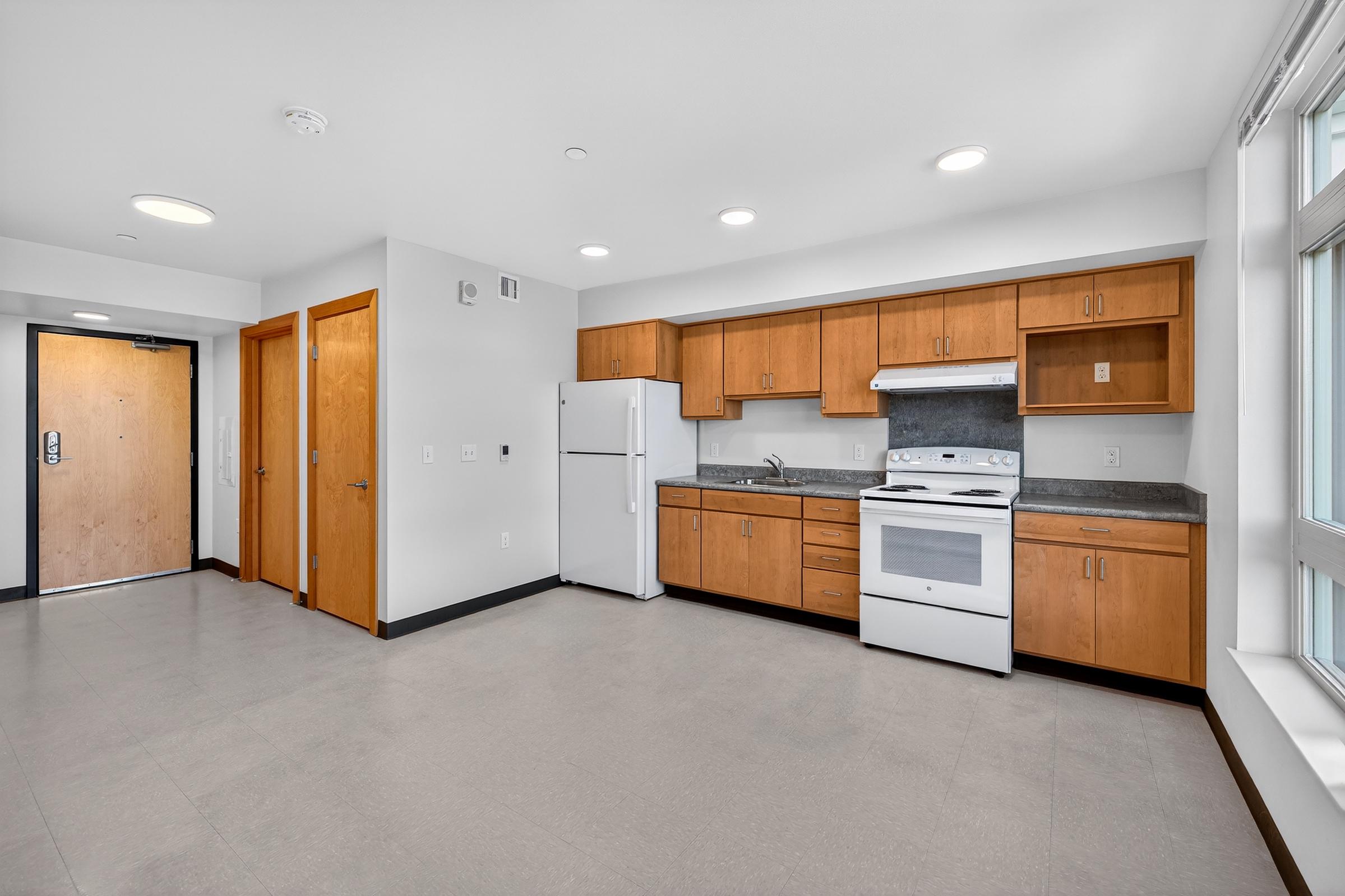 A modern kitchen with wooden cabinets, a white refrigerator, and a white stove. There are gray countertops, and the room features a large window letting in natural light, along with an open layout leading to an entrance door. The floor is tiled in light gray.