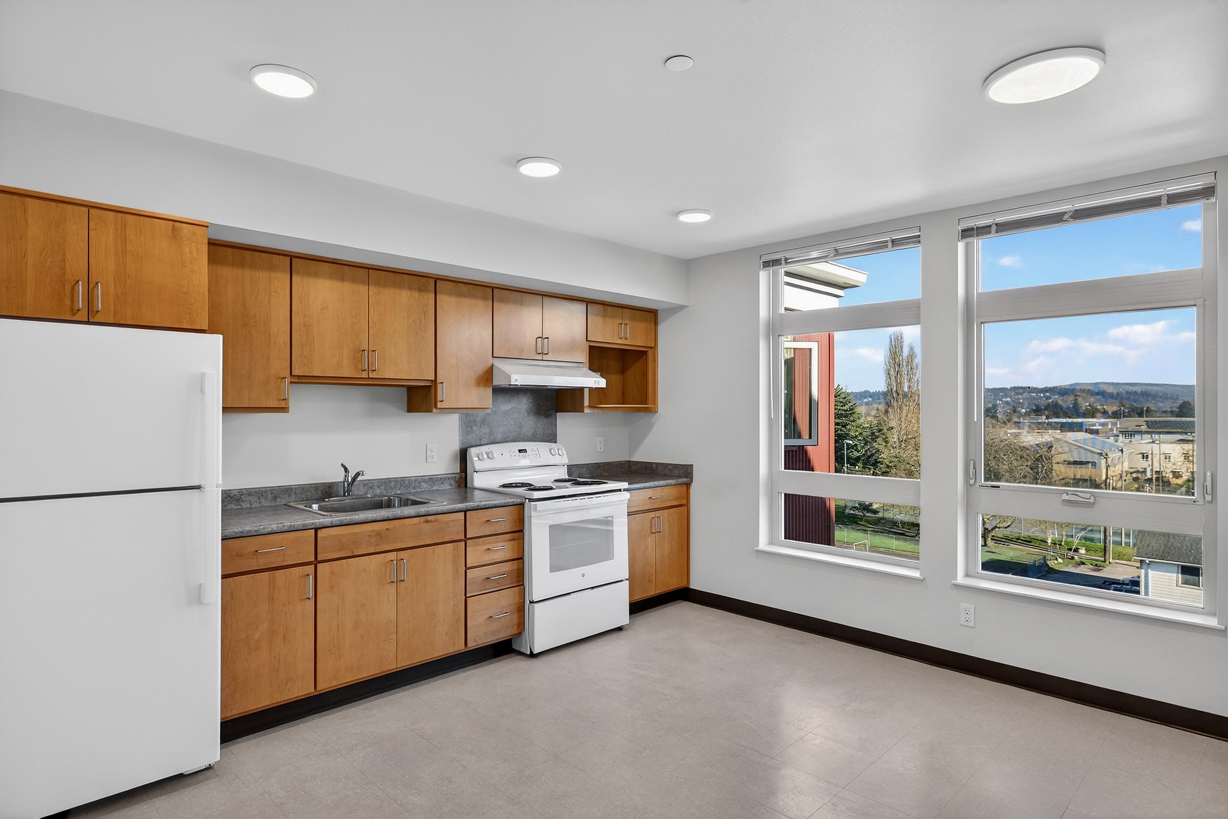 Modern kitchen featuring wooden cabinets, a white stove, and a refrigerator. The countertop is gray, and large windows provide a view of the outside. Bright lighting fixtures illuminate the space, creating a welcoming atmosphere.
