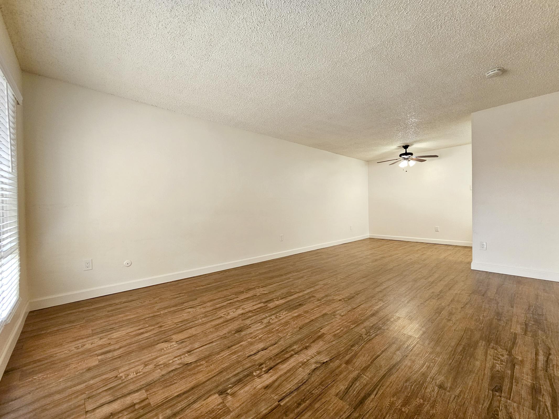 Spacious empty room with light-colored walls and hardwood-style flooring. A ceiling fan is visible in the center, and there are windows allowing natural light to enter. The overall atmosphere is bright and airy, suitable for various interior design possibilities.
