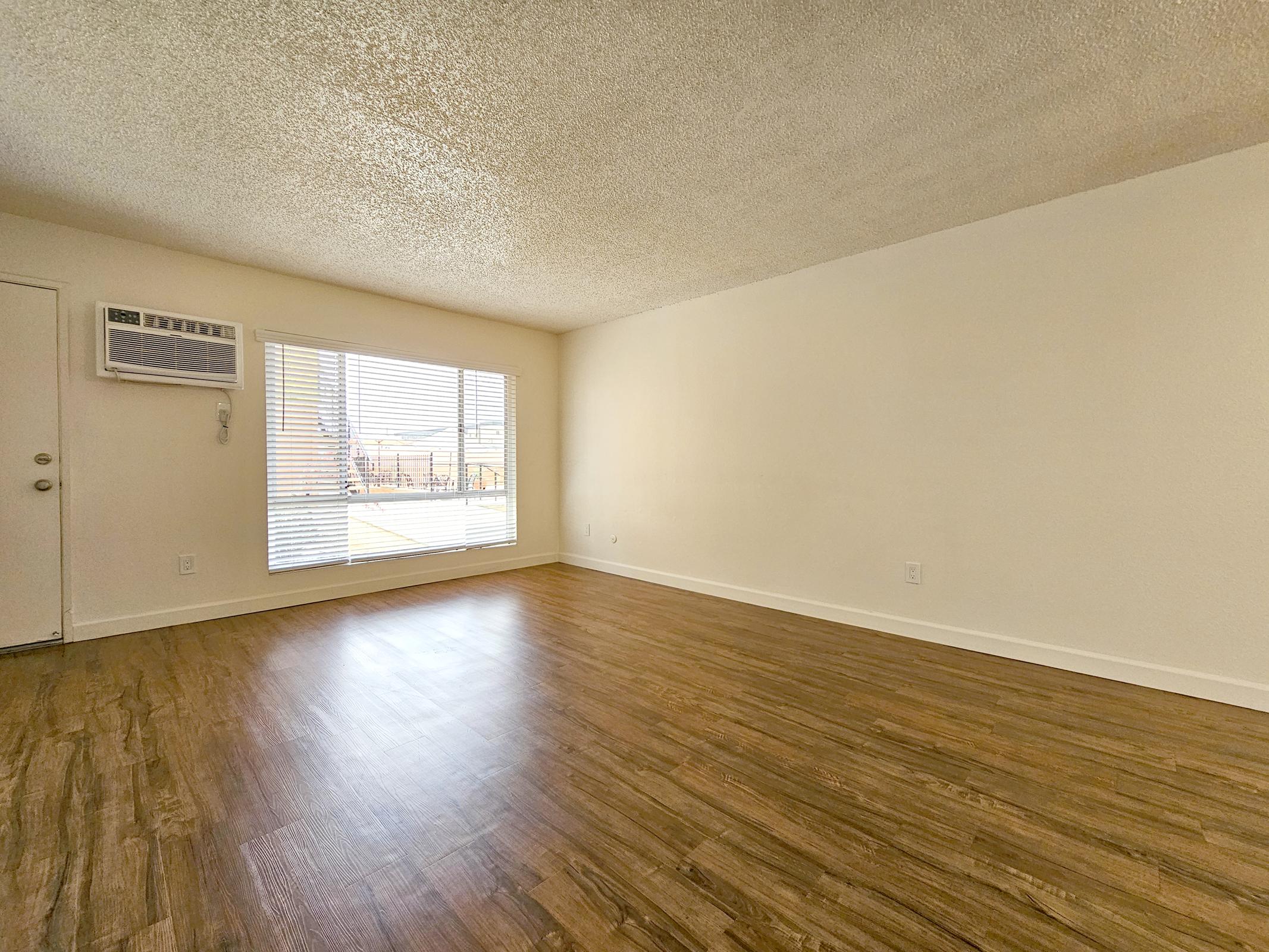 Empty room with light-colored walls and a textured ceiling. A large window with blinds allows natural light to fill the space, revealing a view outside. The floor features warm wood-like laminate flooring. An air conditioning unit is mounted on the wall near the window.