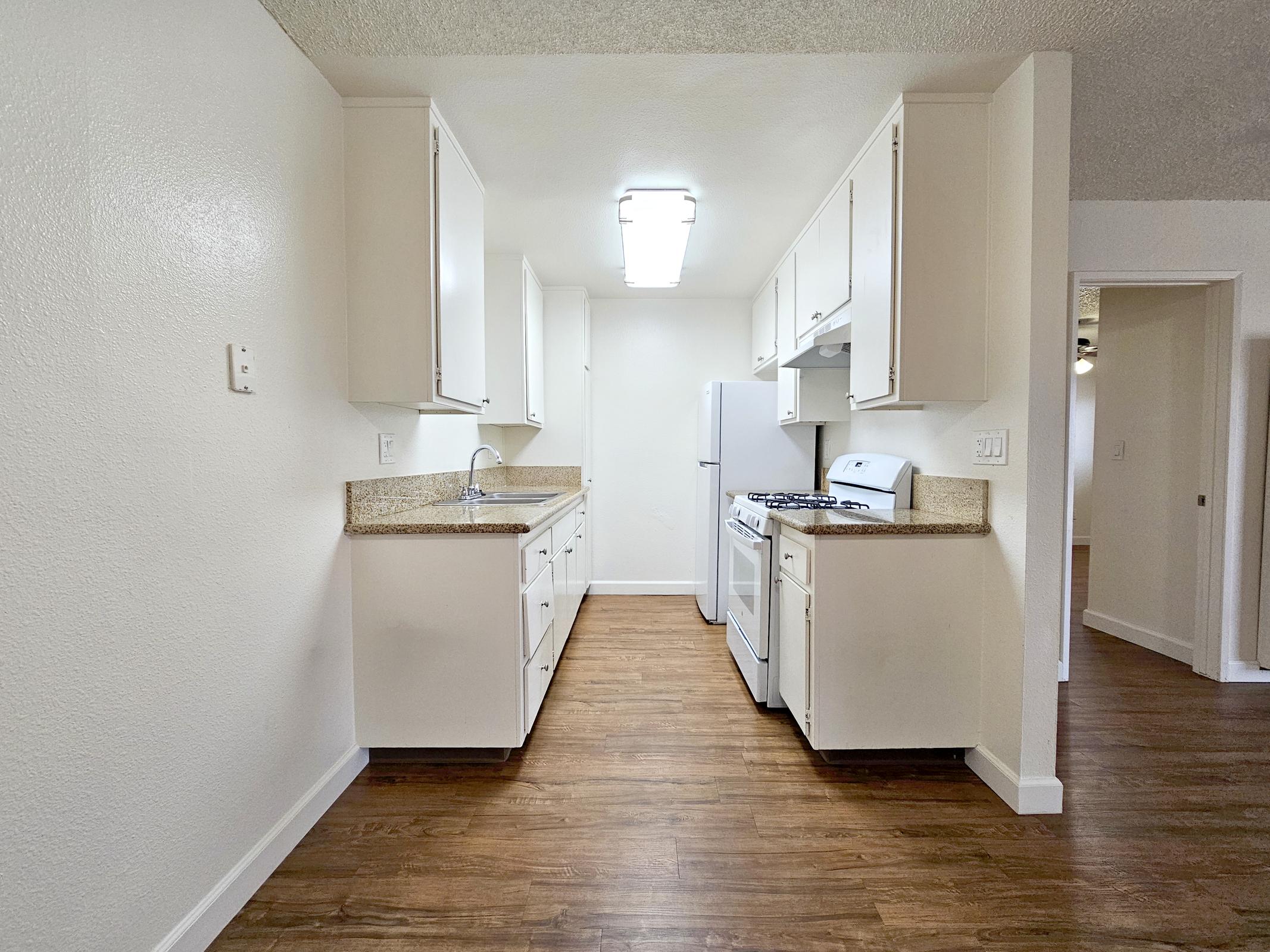 A bright, newly renovated kitchen featuring white cabinets, a stainless steel sink, and a gas stove. The countertops are light-colored, and the flooring is wood-like. There is a refrigerator opposite the stove, with ample storage space and a simple, modern design. The room is well-lit and inviting.