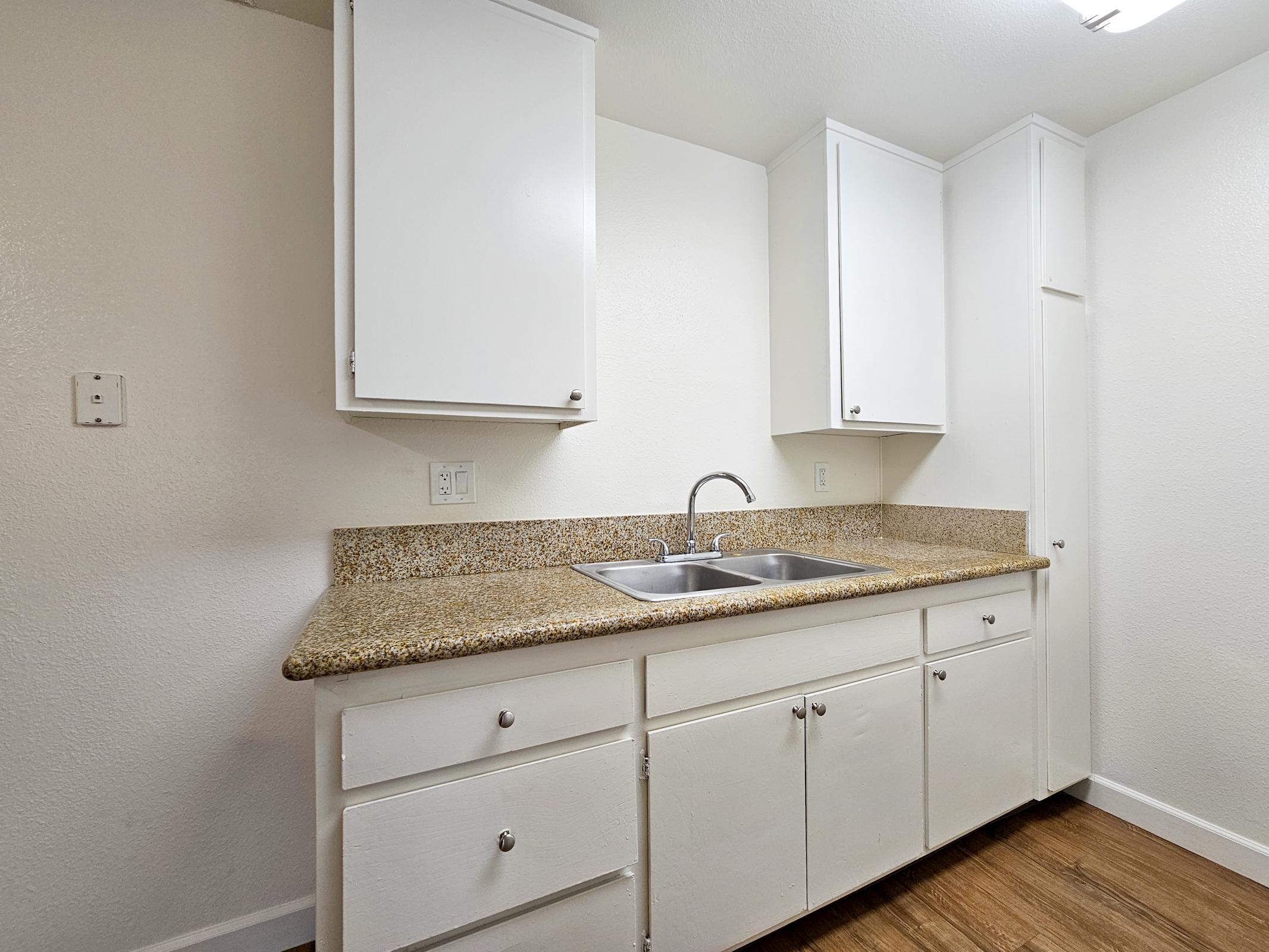 A clean, modern kitchen featuring white cabinets, a granite countertop, a double sink with a faucet, and a light-colored wall. The flooring is made of wood, contributing to the overall bright and spacious feel of the area.