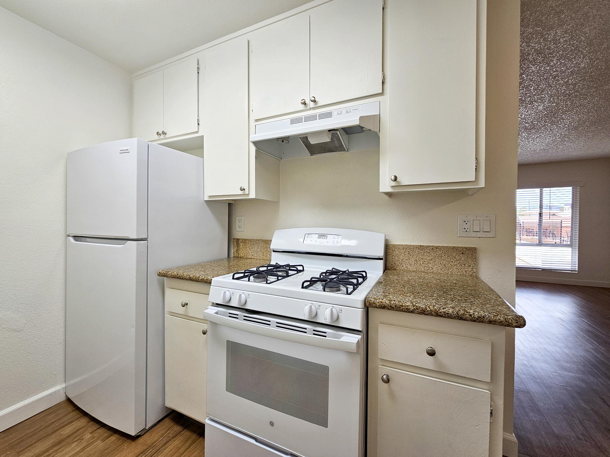 A modern kitchen featuring a white refrigerator, a gas stove with an oven, and a kitchen hood. The cabinets above the countertop are white, and the countertop has a granite finish. A window in the background allows natural light, showcasing a wooden floor.