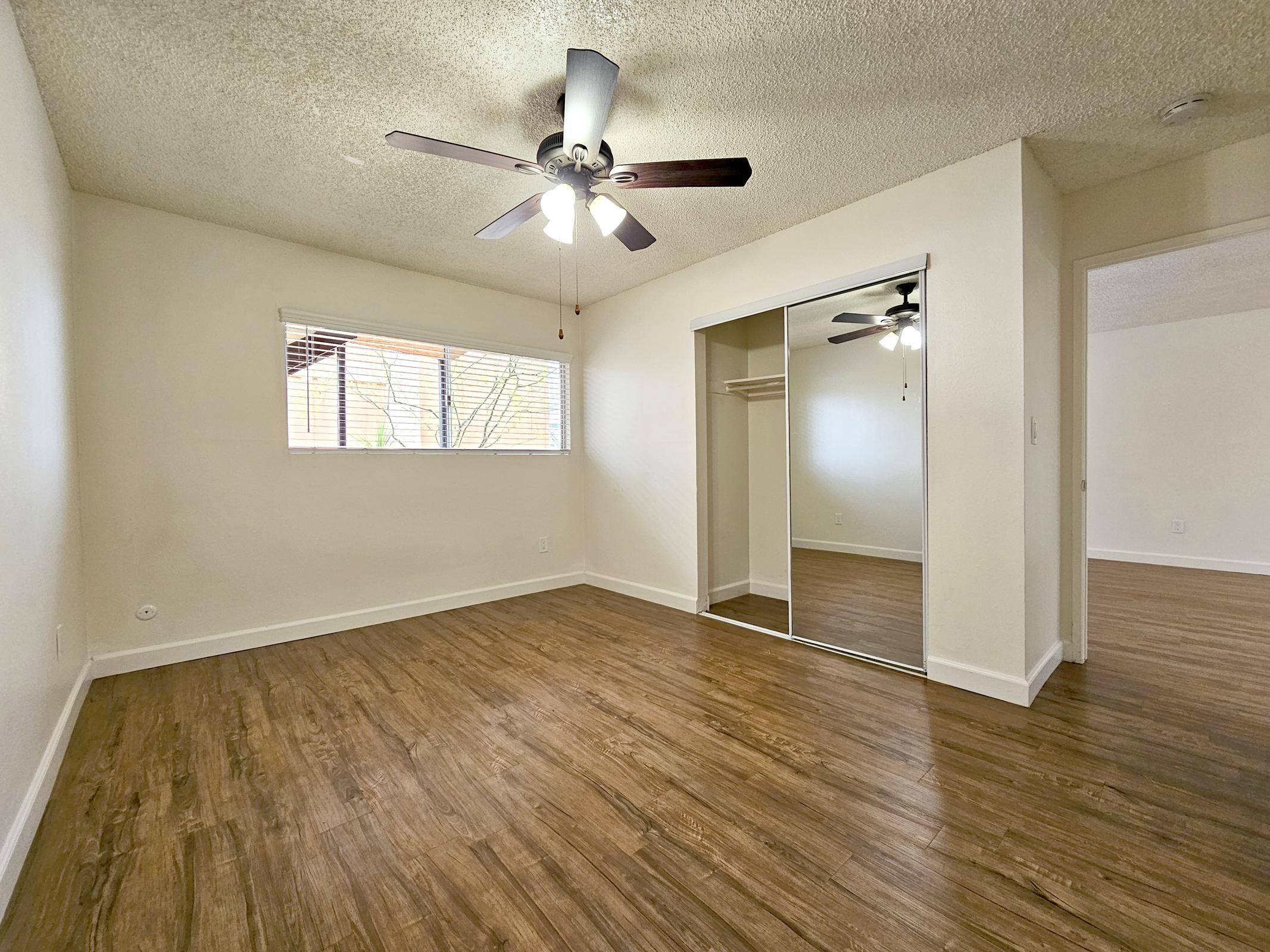 Bright and spacious room with a ceiling fan, featuring wood-like flooring and a large window letting in natural light. A mirrored closet is visible along one wall, showcasing the room's openness. The walls are light-colored, creating a clean and inviting atmosphere.