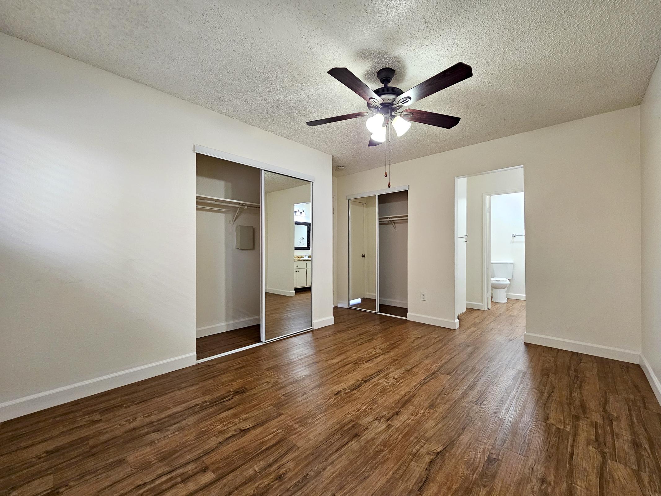 A spacious room with wooden flooring, featuring a ceiling fan and large mirrored closets. There are two doorways leading to an adjoining area, one of which includes a visible bathroom. The walls are painted white, and natural light enters through a window.