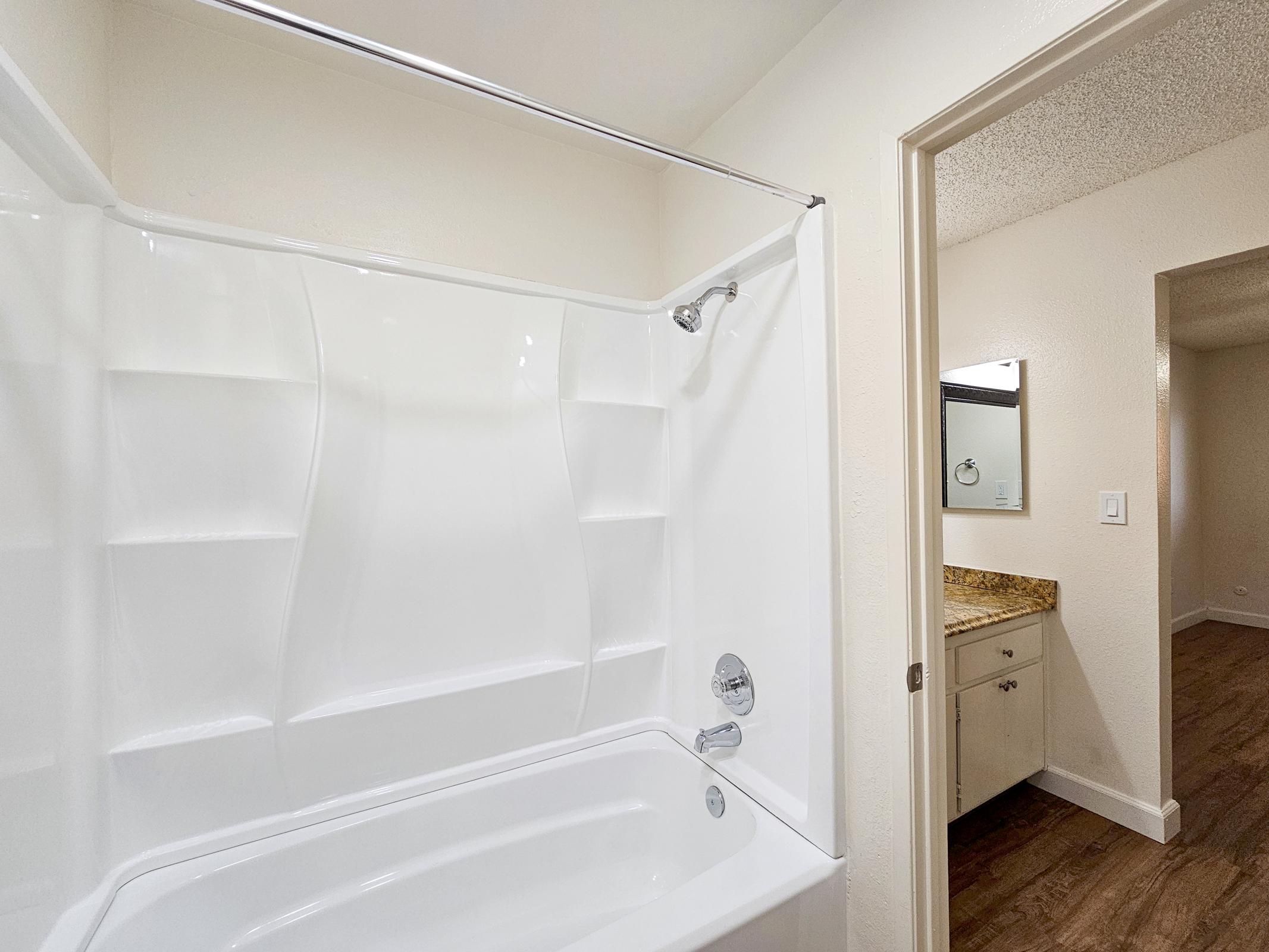 A clean and modern bathroom featuring a white tub and shower combo with a sliding glass door. The light-colored walls create an airy feel. A doorway to the right leads to a hallway with a glimpse of a bathroom vanity sink and mirror in the adjacent room. The floor has a wood-like finish.