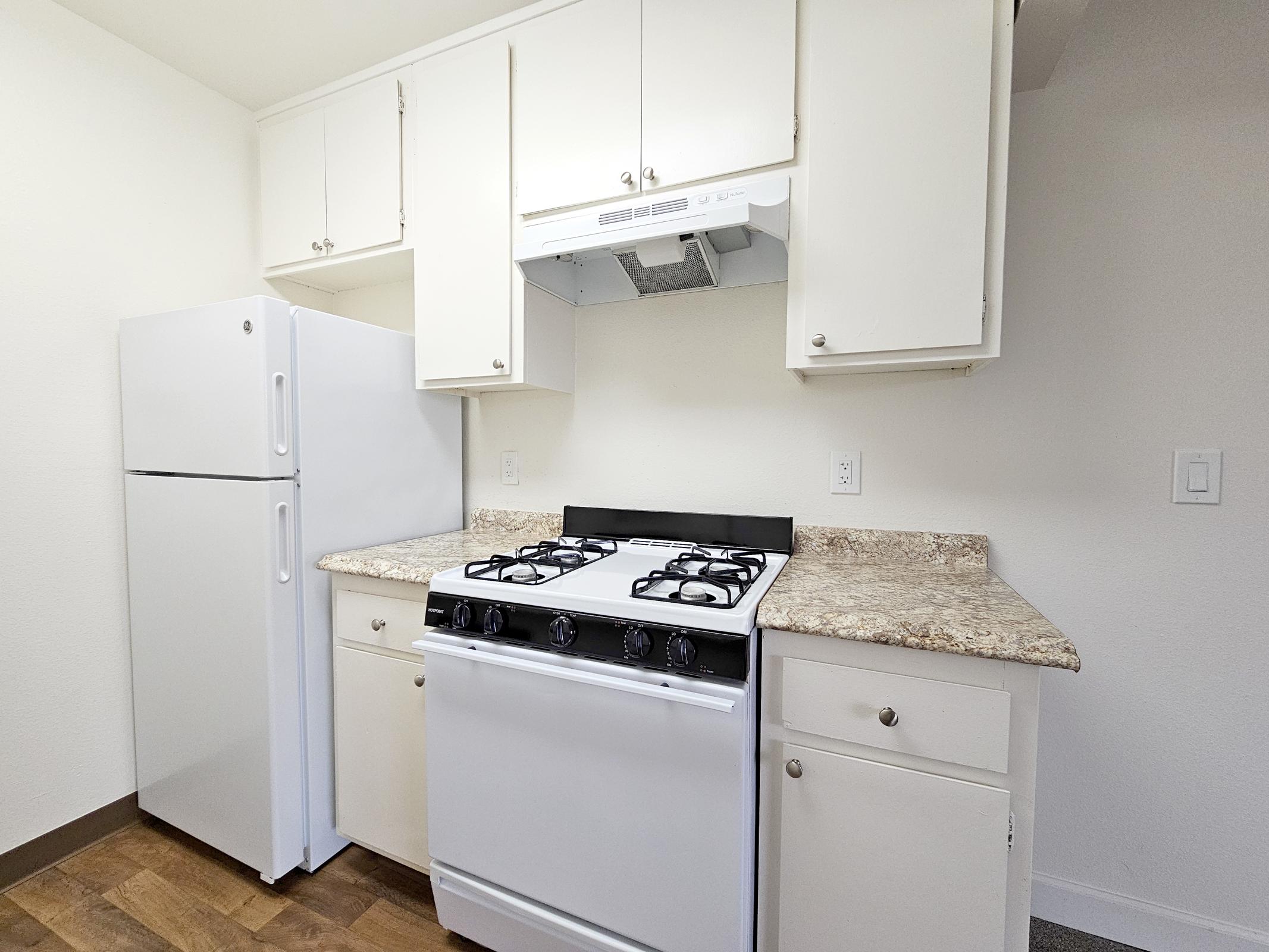 A small kitchen featuring a white refrigerator, a gas stove with a black and stainless steel range hood, and light-colored cabinets. The countertops are a speckled design, and the floor is made of wood. The walls are painted in a light tone, creating a bright and clean look.