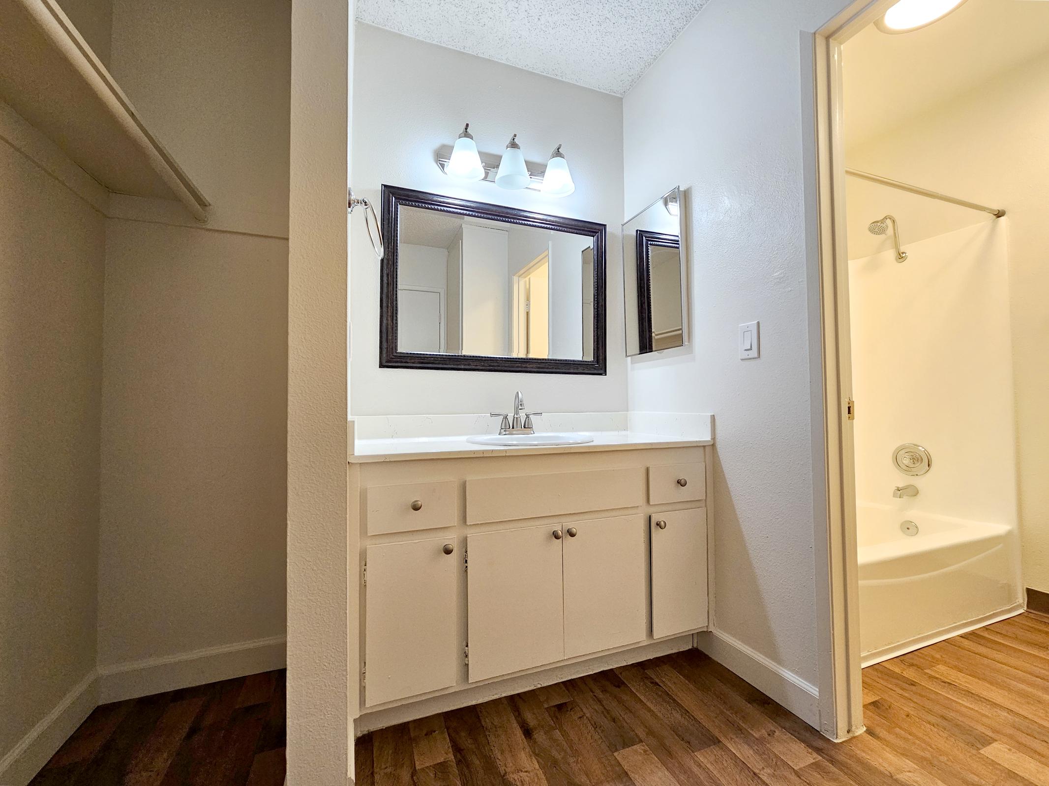 A bathroom featuring a sink with a large mirror above, white cabinetry, and three light fixtures. In the background, there is a shower with a sliding door, and the floor is finished with wood-like laminate. The walls are painted in a neutral color, creating a clean and modern look.