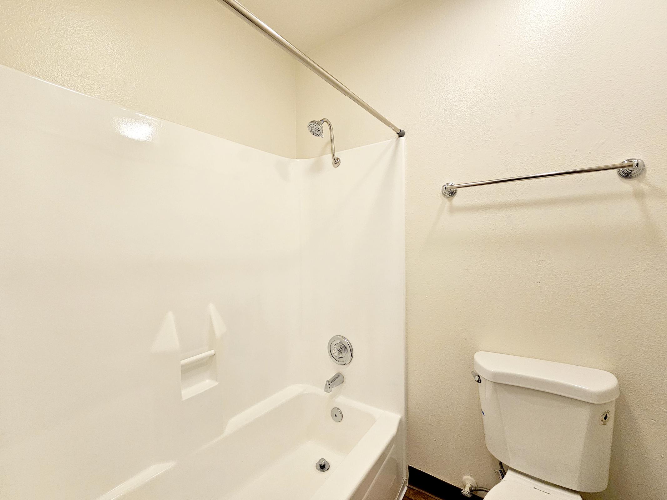 A clean, modern bathroom featuring a white bathtub with a showerhead, a toilet, and a grab bar mounted on the wall. The walls are painted a light color, creating a bright and spacious atmosphere. The flooring is neutral, complementing the bathroom fixtures.