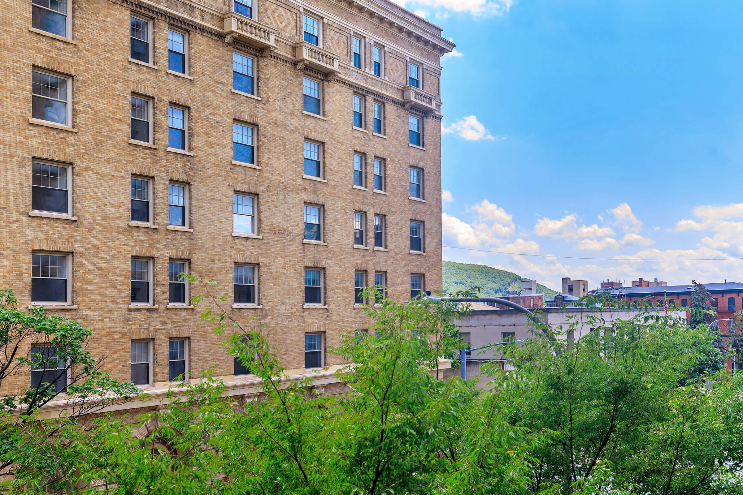 A view of a multi-story brick building with multiple windows, set against a bright sky with scattered clouds. In the foreground, lush green leaves are visible, suggesting a tree or plants nearby. The background features a hillside and urban landscape.