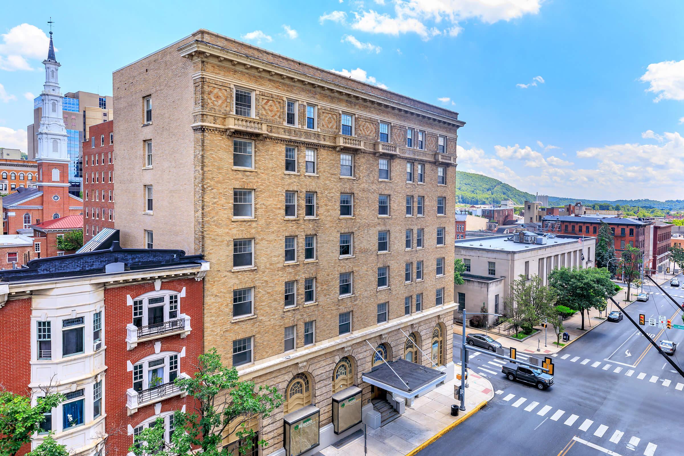 A view of a city street featuring a historic brick building with multiple stories, surrounded by other buildings. In the background, a church steeple rises against a blue sky with clouds, and a hillside is visible. The scene captures urban architecture and a vibrant streetscape.