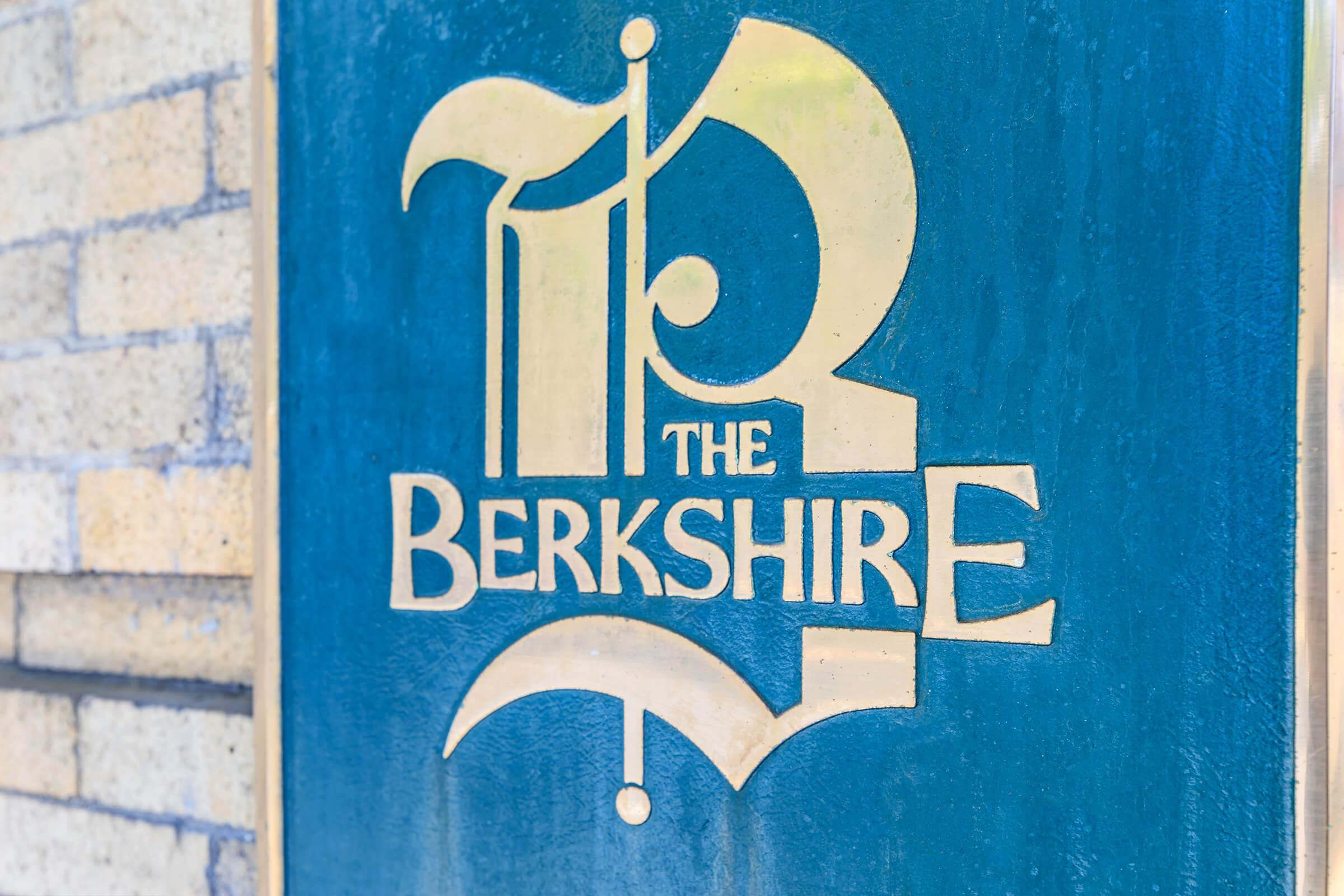 Sign featuring the text "The Berkshire" in a stylish font, with a decorative design above the name. The sign has a blue background and golden lettering, set against a brick wall.