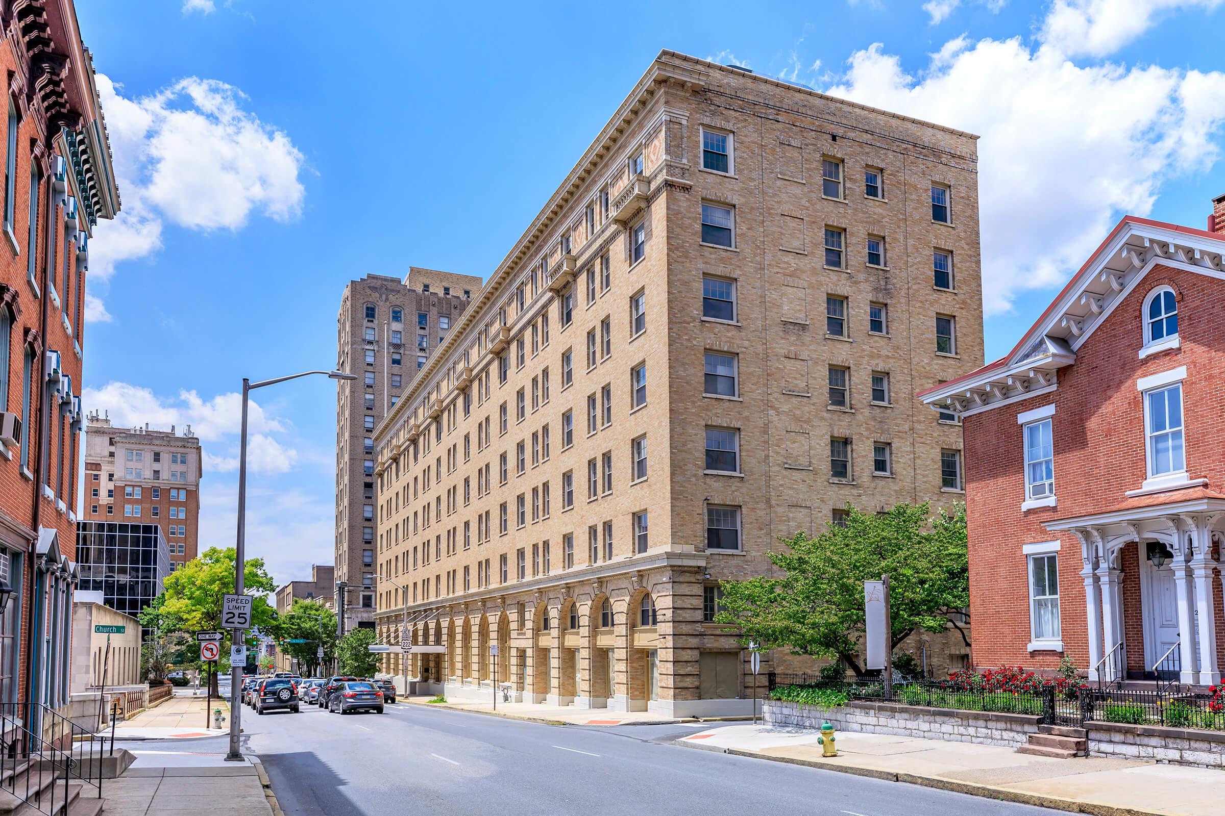 A view of a city street featuring a tall, historic brick building with large windows, flanked by modern and traditional architecture. The sky is bright with fluffy clouds, and trees line the street. Cars are parked along the road, and a speed limit sign is visible.