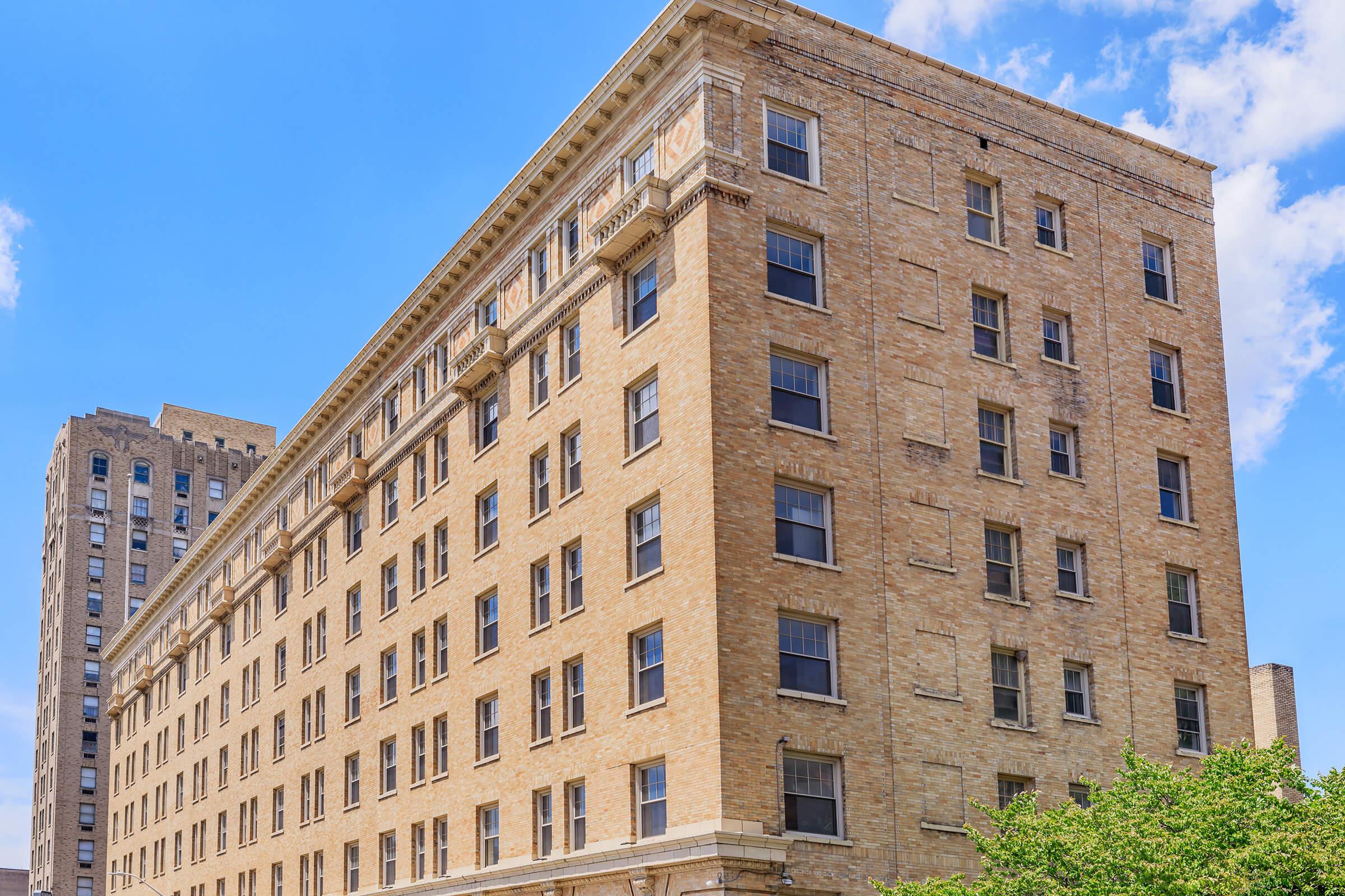 Brown brick apartment building with multiple windows and a detailed cornice, set against a blue sky with scattered clouds. Additional buildings are visible in the background, contributing to an urban landscape. Green foliage is present at the base.