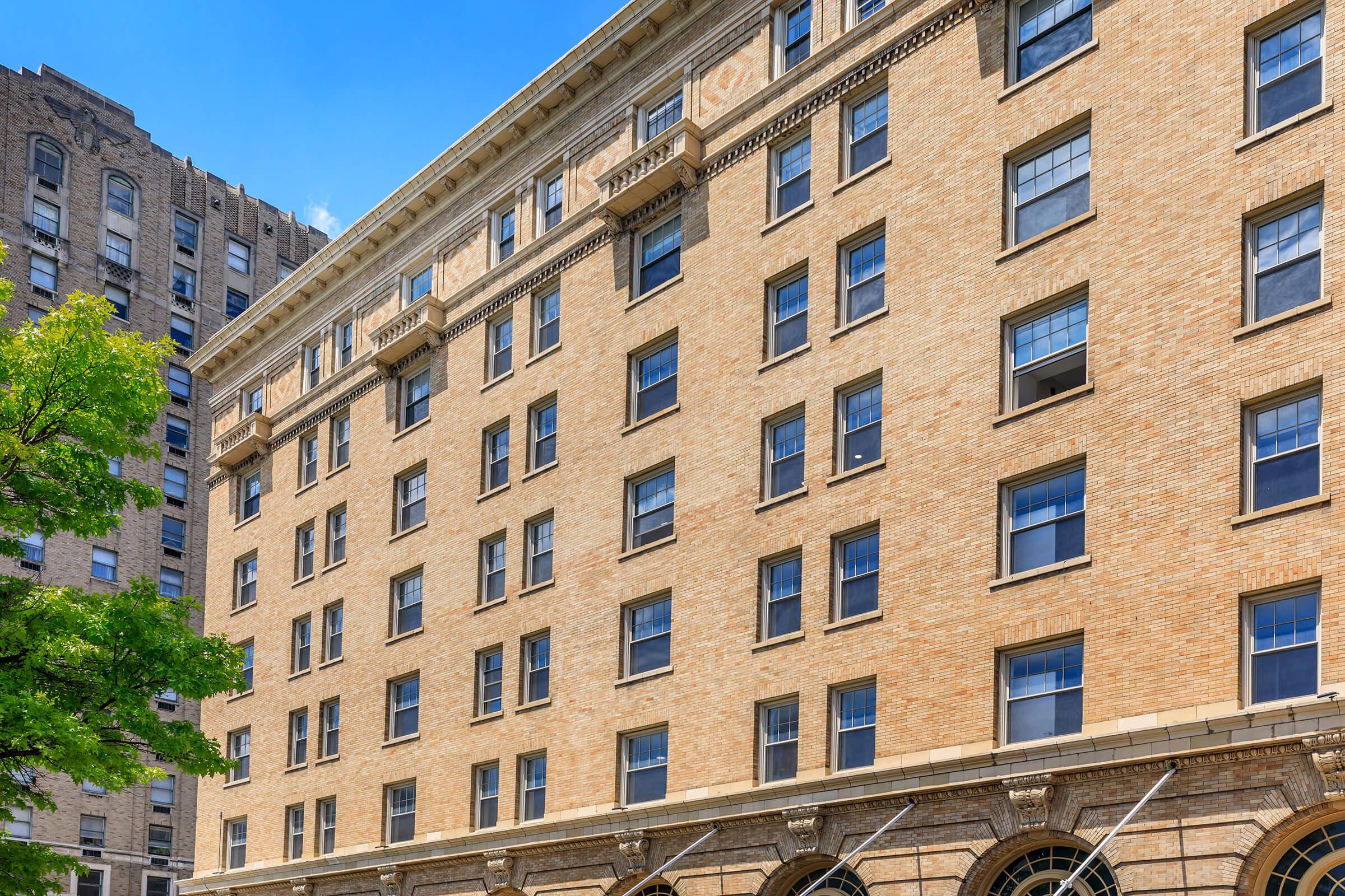A close-up view of a historic brick building featuring multiple rectangular windows with decorative stone accents. The facade is well-maintained, showcasing classic architectural details, and is surrounded by lush green trees under a clear blue sky.