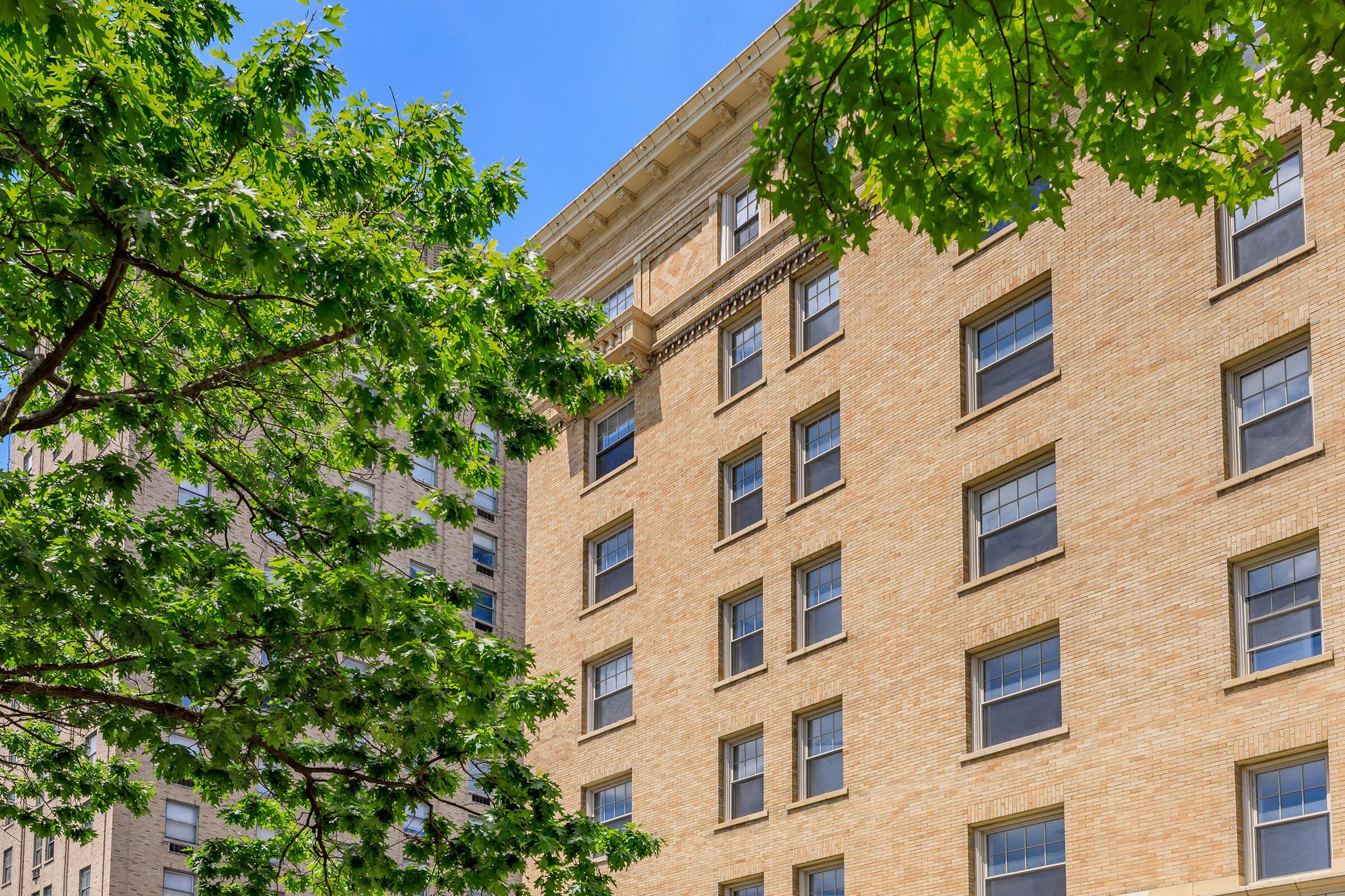 A close-up view of a brick building with multiple windows, framed by green leaves from nearby trees against a clear blue sky. The architectural details highlight the building's structure and the natural surroundings.