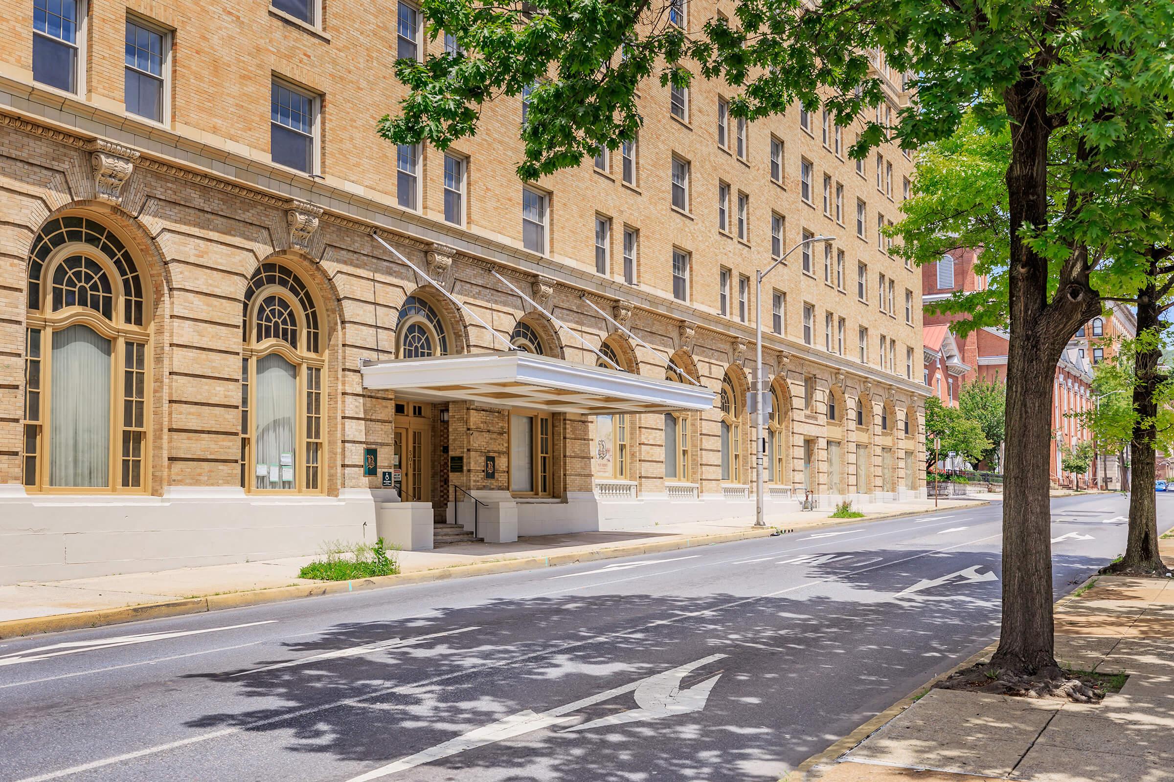 A view of a historic building with a brick facade and arched windows, featuring a covered entrance on the side. There is a tree along the sidewalk, and the scene is set on a quiet street with clear skies.