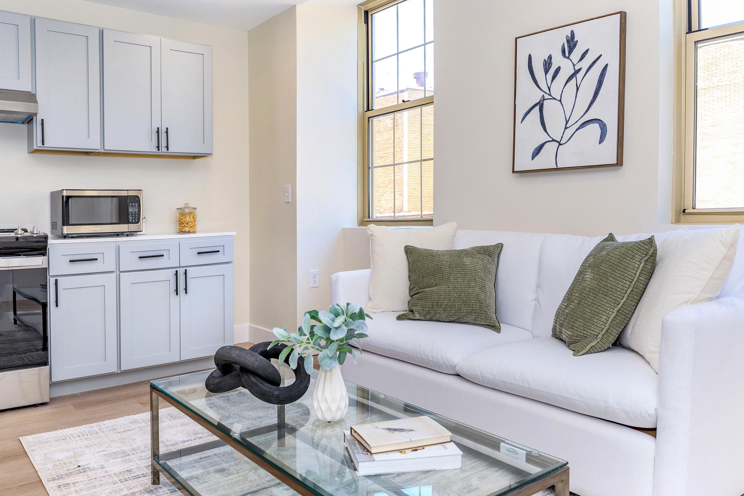A modern living room with a white sofa adorned with green and white pillows, a glass coffee table featuring a decorative vase and a book. The kitchen area in the background includes light gray cabinets, a microwave, and a window letting in natural light. A framed botanical print hangs on the wall.