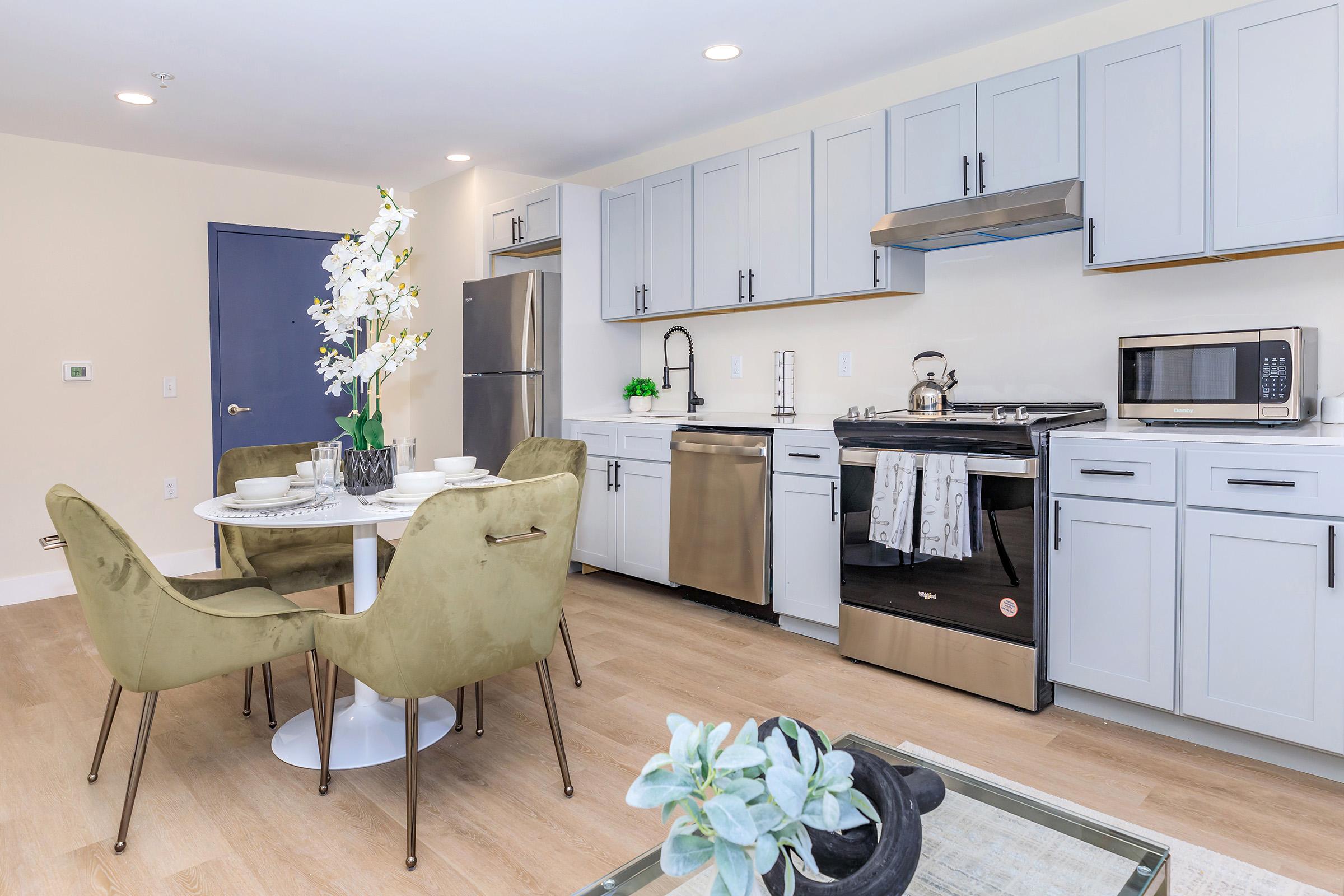 A modern kitchen featuring light gray cabinets, stainless steel appliances, and a dining area. A round white table is set with dishes and surrounded by green upholstered chairs. Decorative flowers add a touch of elegance, while a small plant is visible in the foreground. Bright lighting enhances the clean and contemporary design.