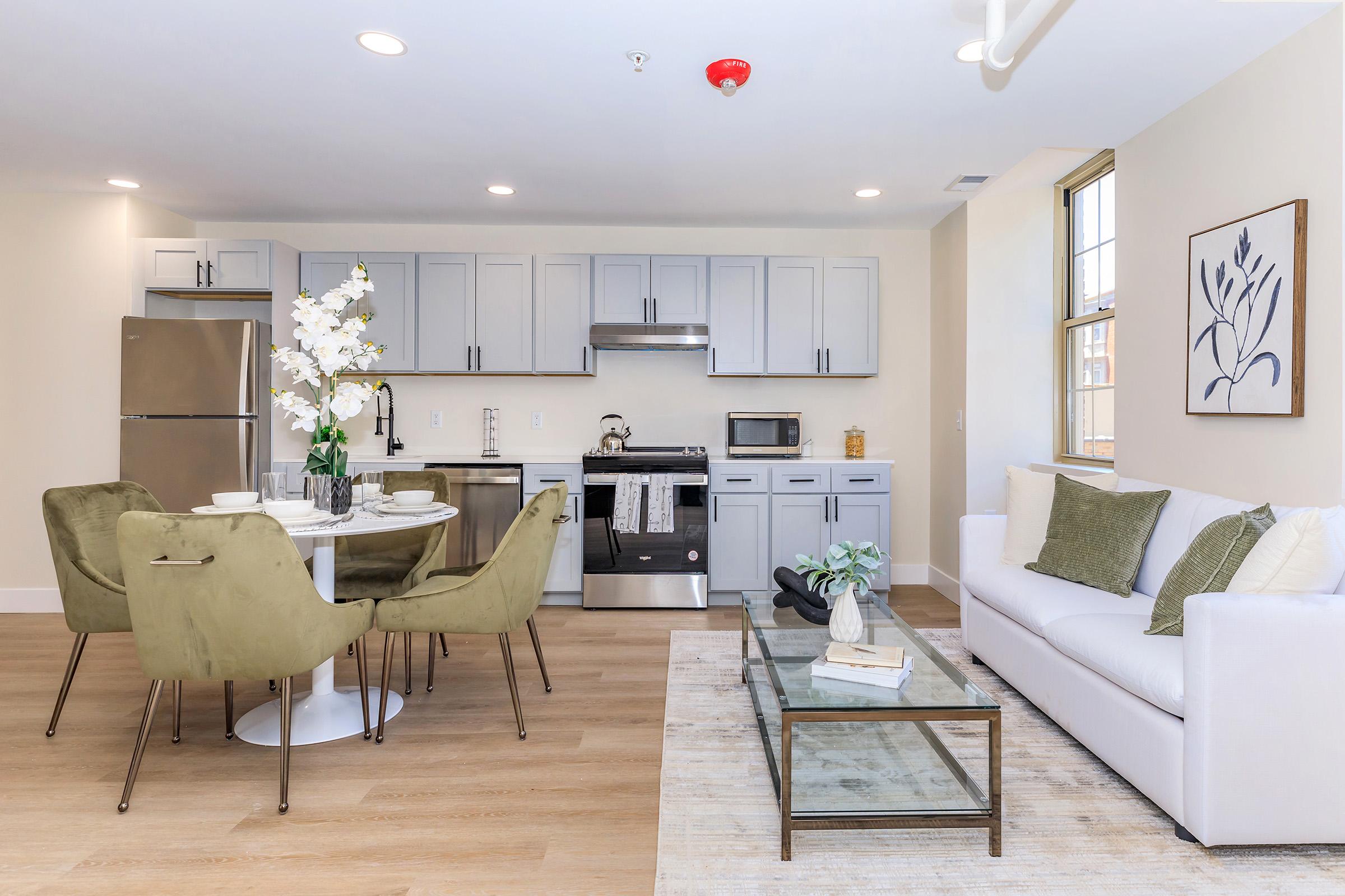 Modern kitchen and living area featuring a round dining table with four green chairs, a sleek kitchen with stainless steel appliances, and a comfortable white sofa. Decor includes a vase with white flowers and a framed botanical print on the wall, set in a bright, open space.