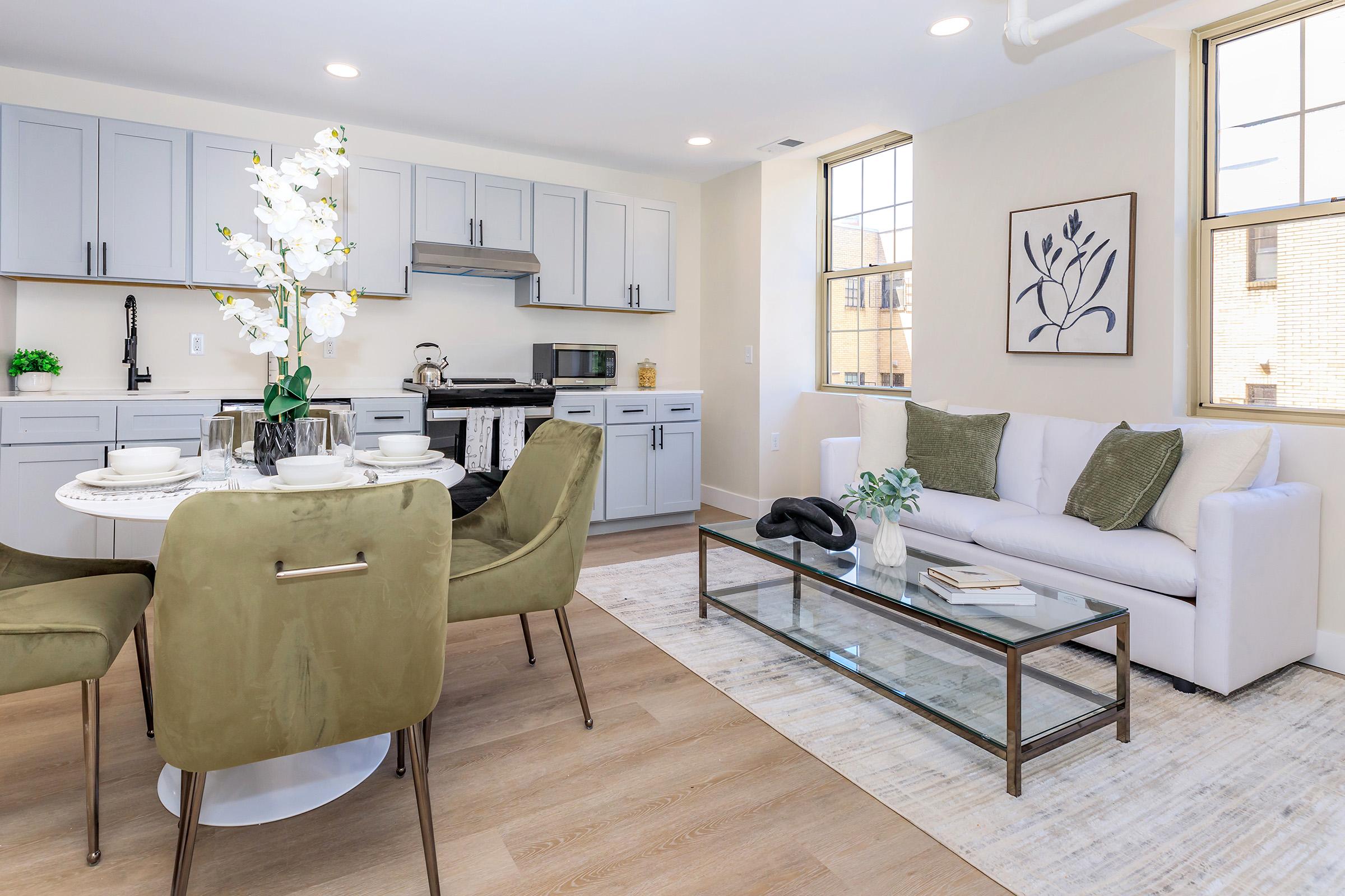 Modern kitchen and living area featuring a light gray cabinetry with a stainless steel range, a white dining table set with dishes, and stylish green chairs. A white sofa with decorative pillows sits beside a glass coffee table on a light area rug, with large windows allowing natural light. A decorative plant is on the table.