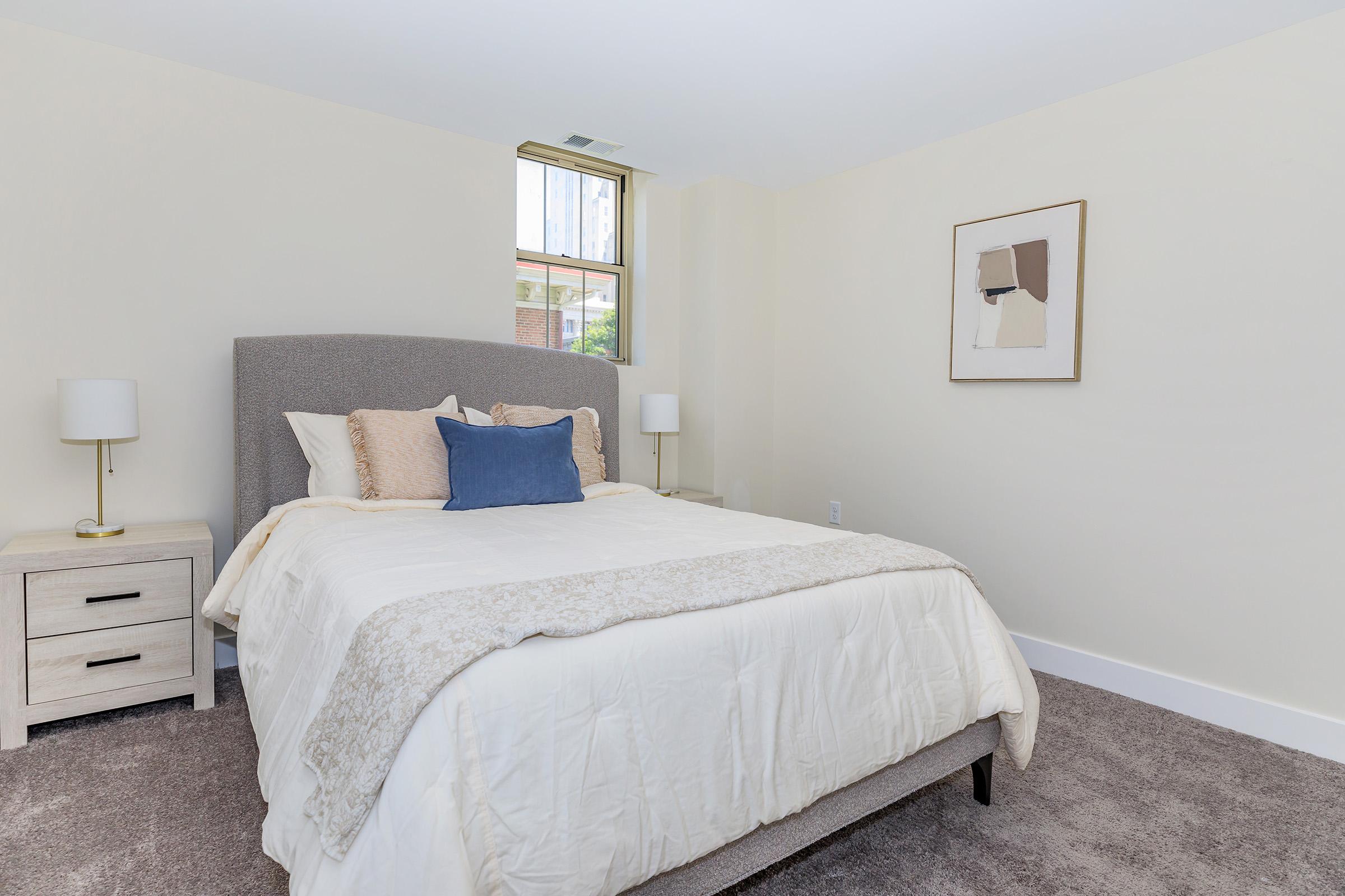 A cozy, well-lit bedroom featuring a light-colored bed with decorative pillows, a gray upholstered headboard, and a beige knitted throw. Adjacent is a nightstand with two table lamps. A framed abstract artwork hangs on the wall, and a small window allows natural light to enter the space. The carpet is a soft, neutral color.