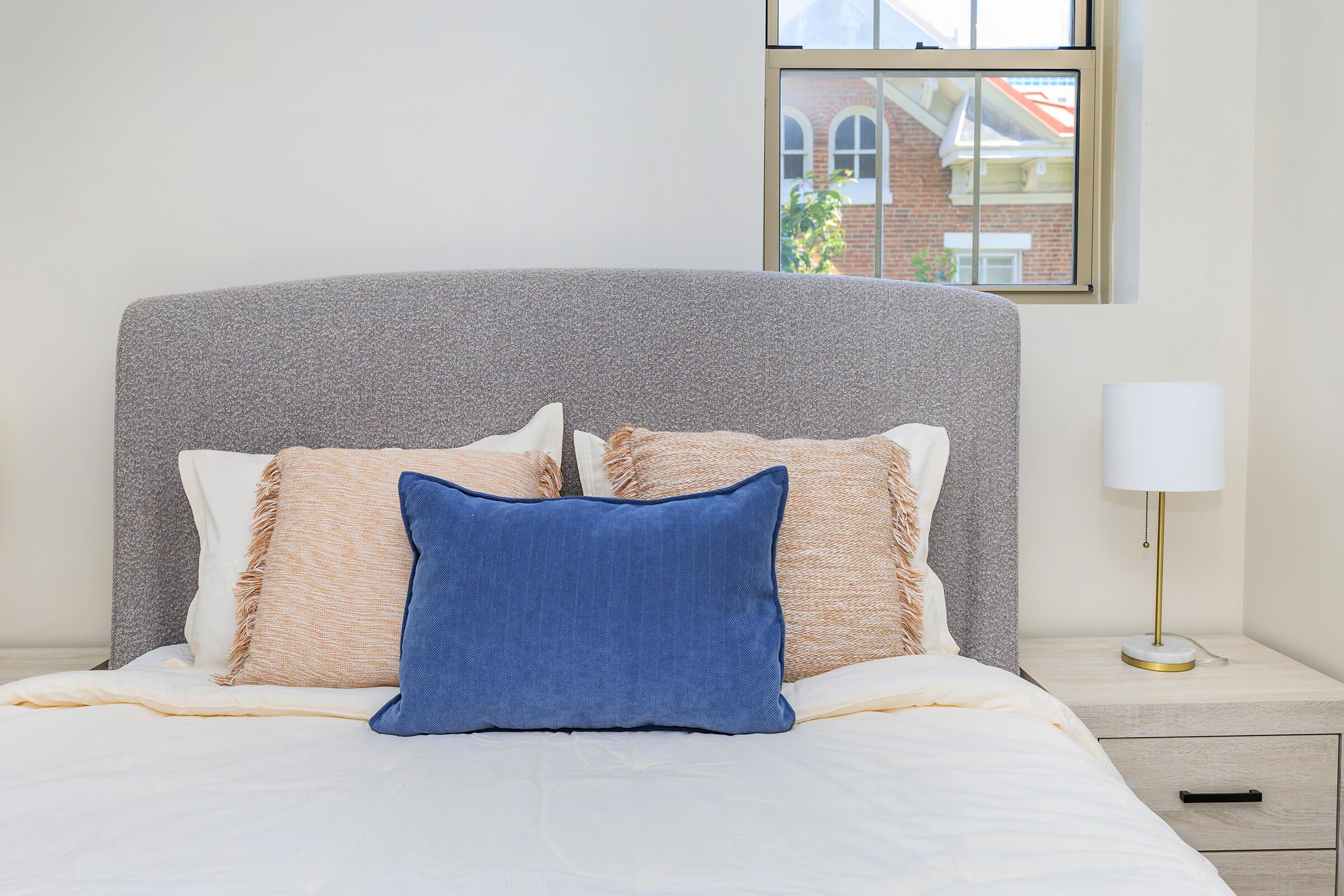 A neatly made bed with a light-colored comforter, featuring several decorative pillows: two cream-colored pillows with textured fringes and a bold blue rectangular pillow. A small lamp sits on a bedside table next to the bed, with a window in the background showing part of a building and greenery outside.