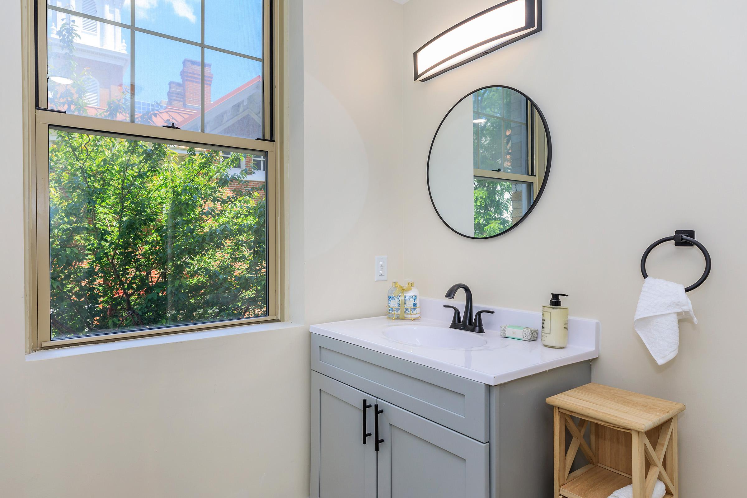 A well-lit bathroom featuring a gray vanity with a white sink, a round mirror above it, a black faucet, and a towel holder. There's a wooden stool beside the vanity. The window shows greenery outside, adding natural light to the room.