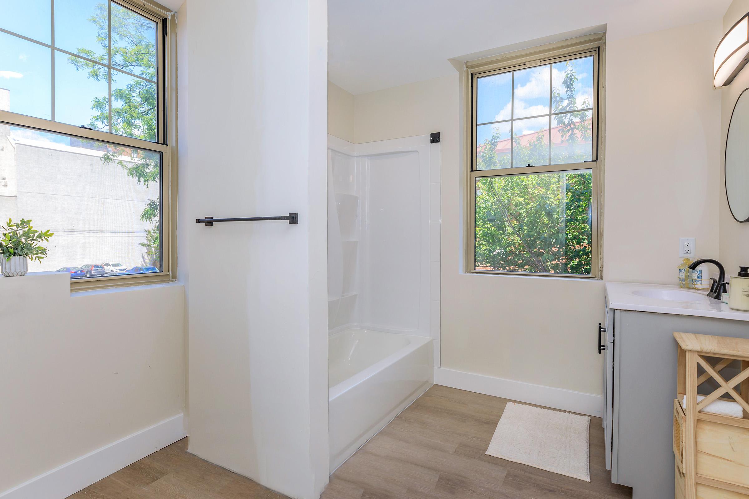 A bright and modern bathroom featuring a bathtub/shower combination, a white vanity with a sink, and large windows letting in natural light. The walls are painted in a soft neutral color, and there is light-colored flooring. A small rug is placed in front of the vanity for added comfort.