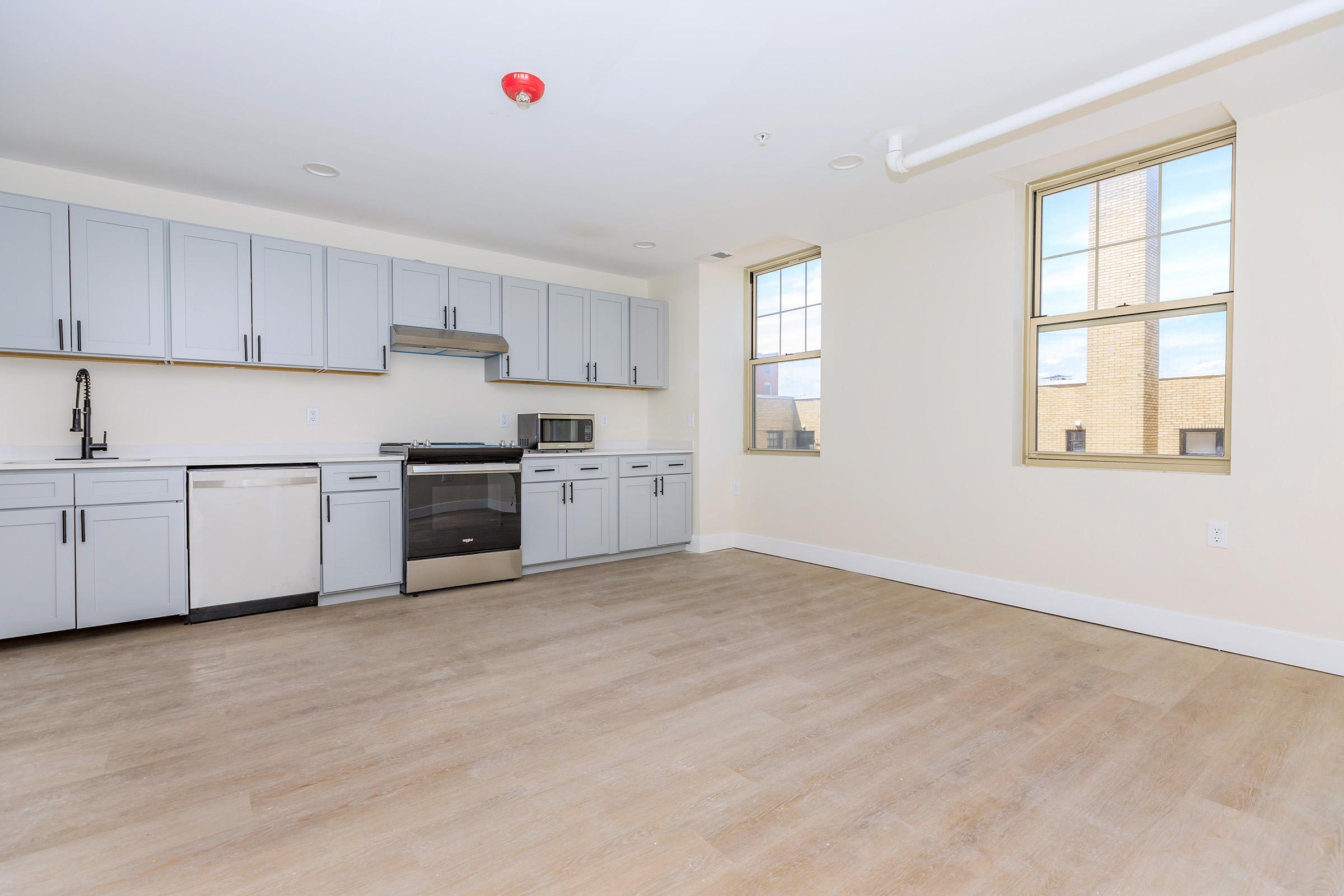 Spacious, modern kitchen and living area featuring light gray cabinets, stainless steel appliances, and large windows allowing natural light. The floor is light wood, creating a warm ambiance in a bright, open layout. Ideal for a contemporary living space.