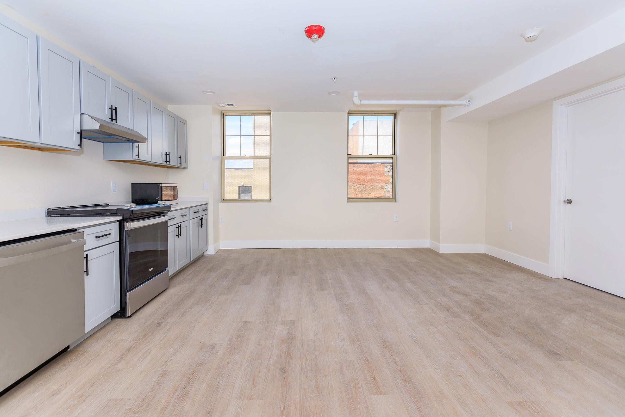A modern kitchen and living space featuring light gray cabinets, stainless steel appliances, a microwave, and a spacious layout with large windows allowing natural light. The flooring is light wood, and the walls are painted in a neutral color. There is a fire alarm on the ceiling.