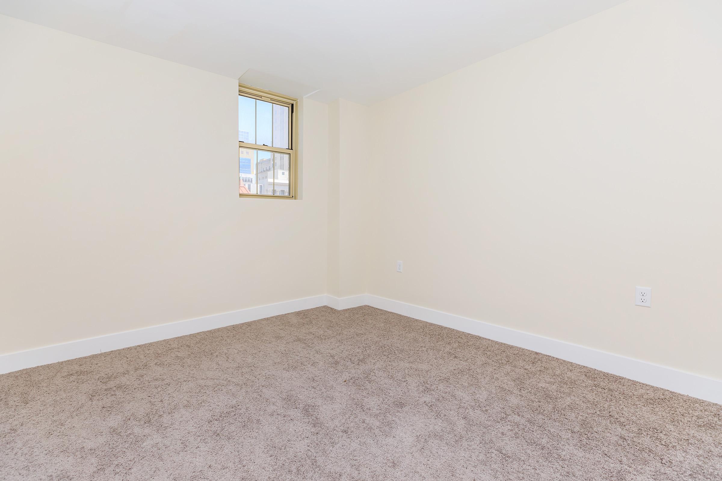 Empty room with light beige walls, a window allowing natural light in, and a soft brown carpeted floor. There are no furniture or decorations present, creating a spacious and minimalistic appearance.