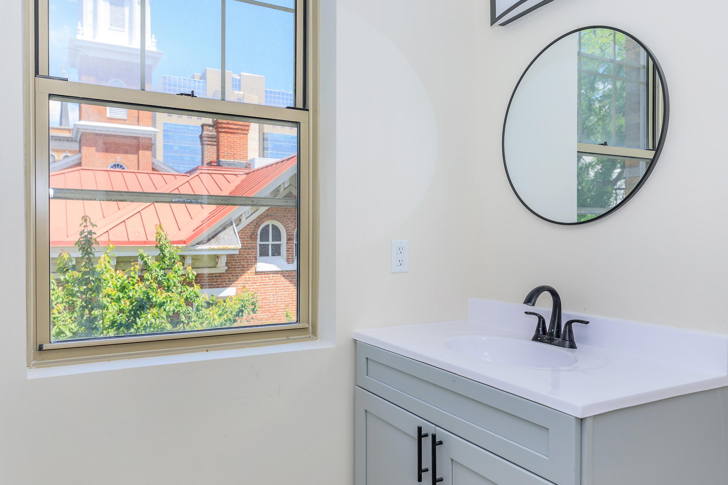 A bright bathroom space featuring a sleek white sink and dark faucet on a light gray vanity. A round mirror hangs above the sink, reflecting natural light from a large window. Outside the window, a view of a neighboring building with a red roof and greenery can be seen.