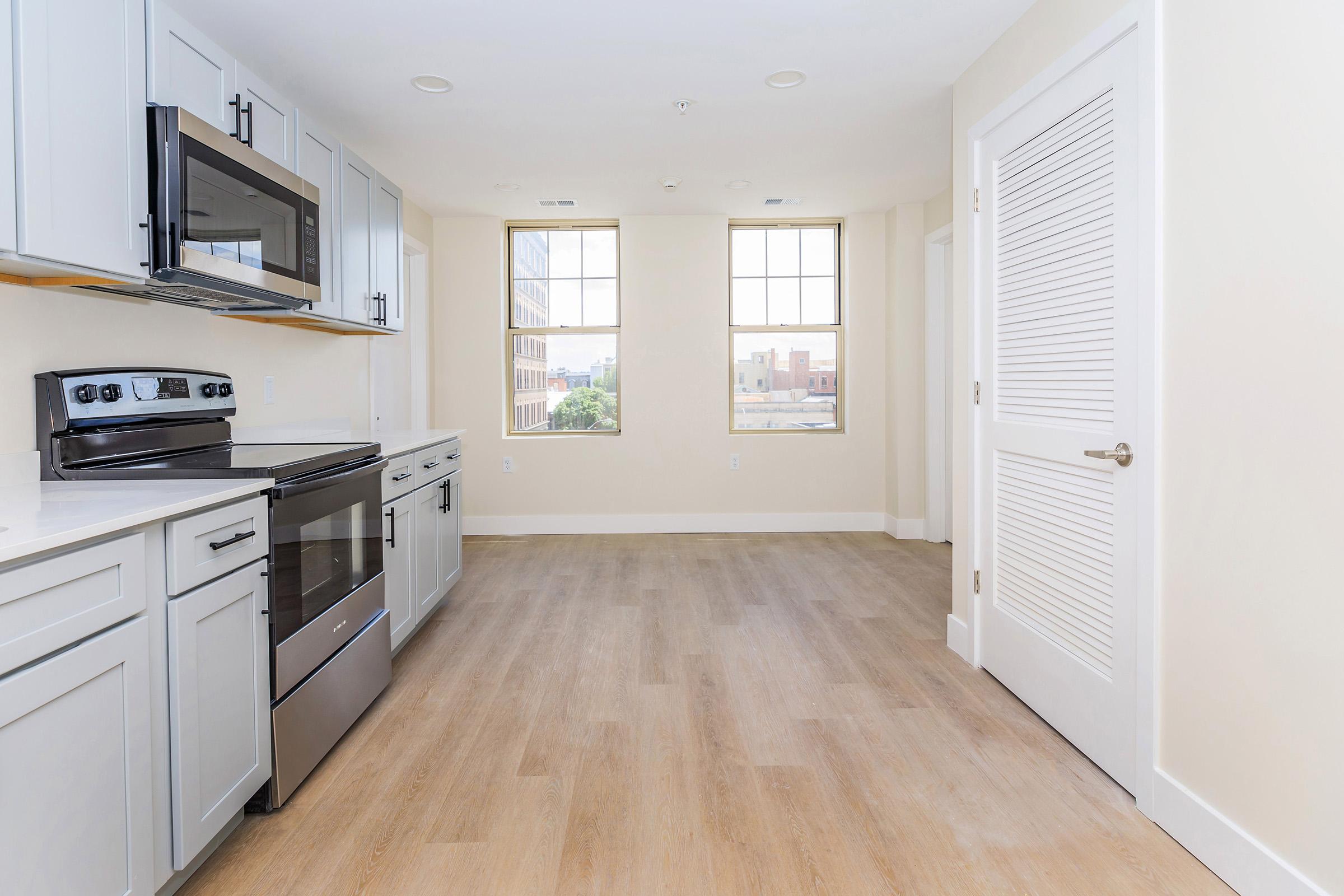 A modern kitchen featuring stainless steel appliances, including an oven and microwave, with light-colored cabinetry. The room has large windows allowing natural light, and a light wood floor. A door on the right leads to another room. The overall design is sleek and contemporary.