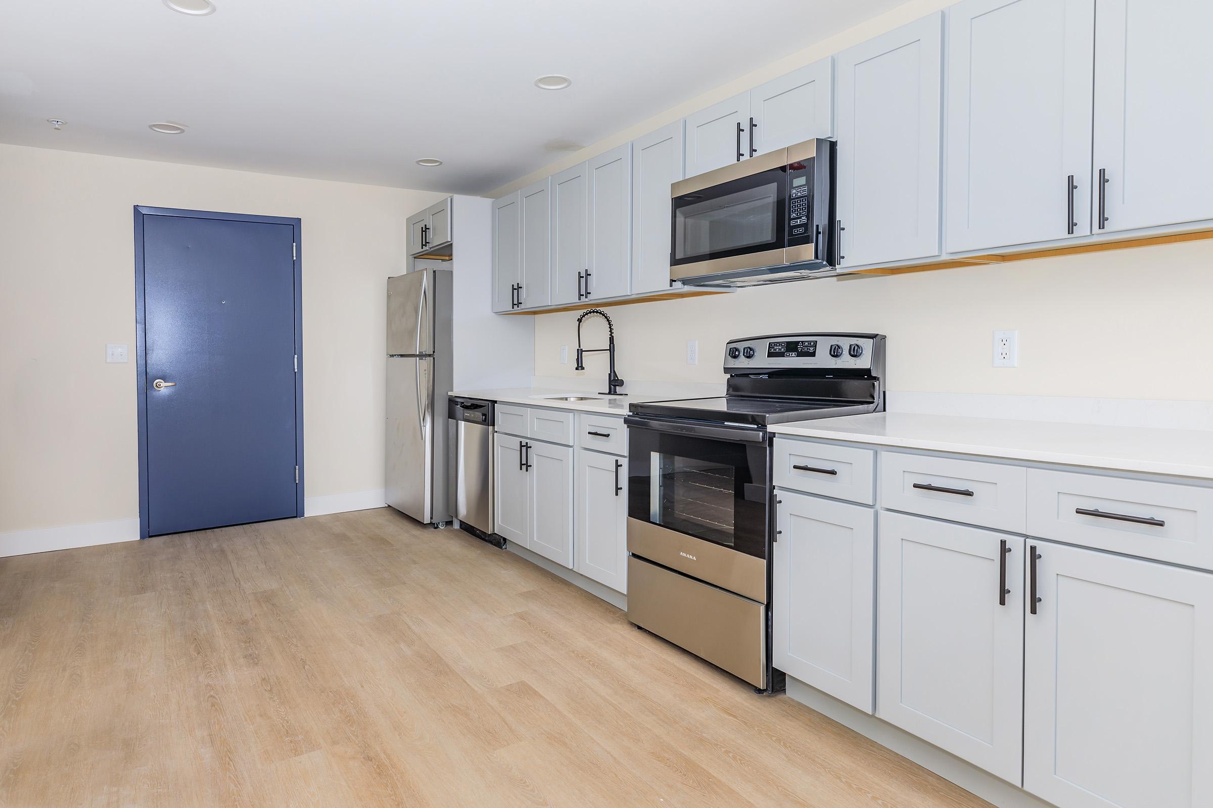 Modern kitchen featuring light gray cabinets, stainless steel appliances including a refrigerator, oven, and microwave, and a neutral-colored wall. The space has wooden flooring and a contemporary faucet above a sink. A blue door is visible on the left side of the image.