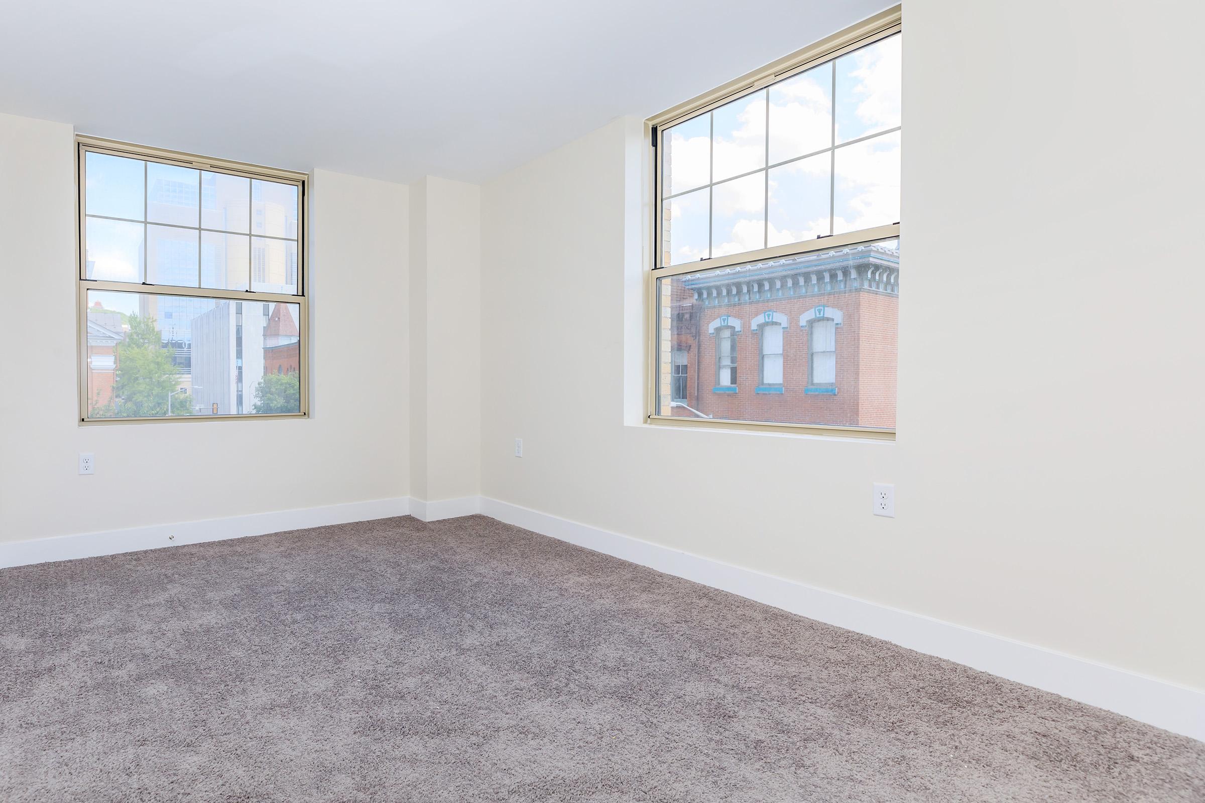 An empty room with light-colored walls and plush carpeting. Two large windows allow natural light to enter, revealing a view of brick buildings outside. The space is clean and uncluttered, suggesting it is newly painted or renovated.