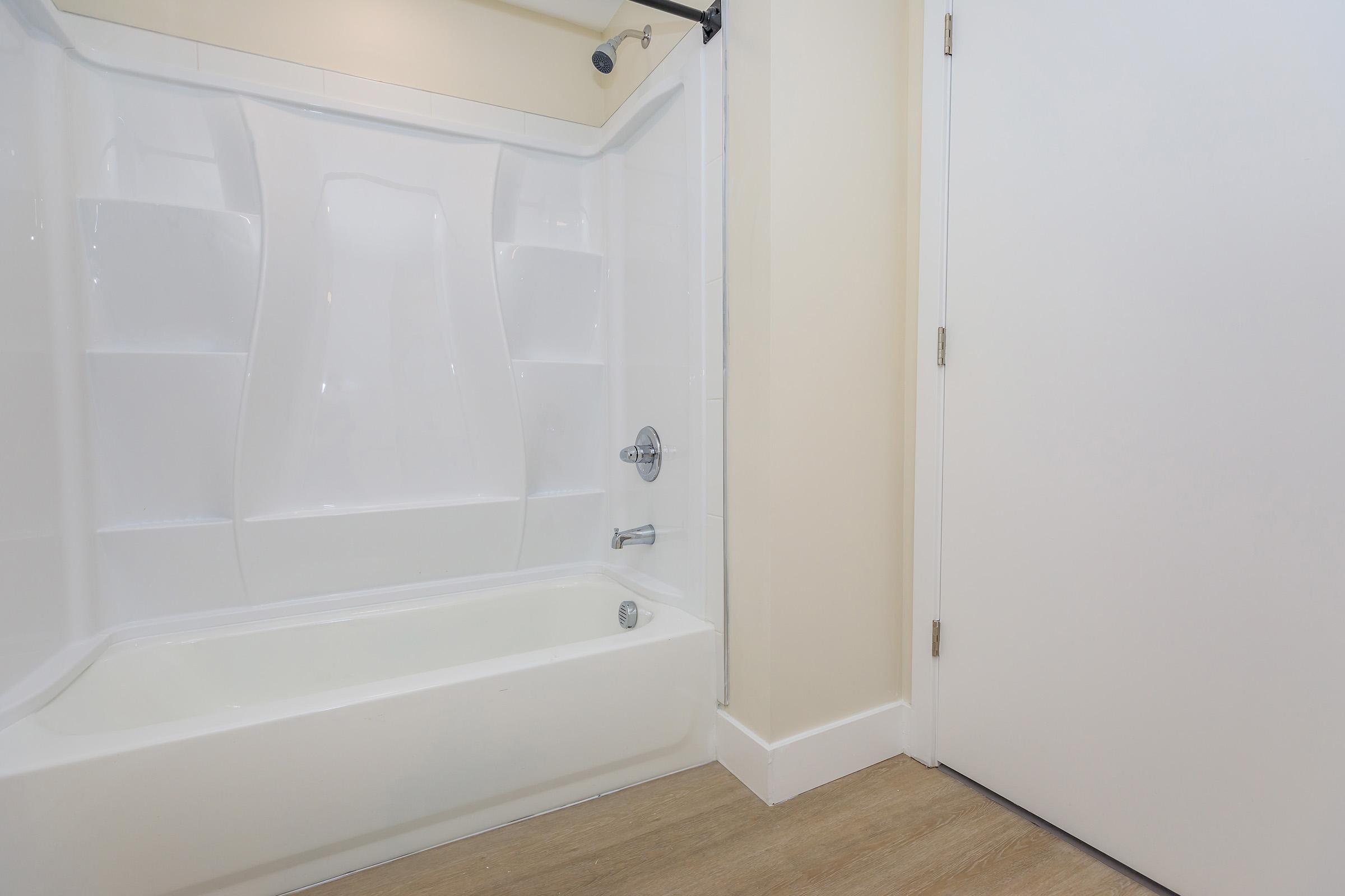 A clean bathroom with a white bathtub and shower enclosure. The wall is painted a light color, and there's a white door partially visible to the right. The floor is finished with a light wooden texture.