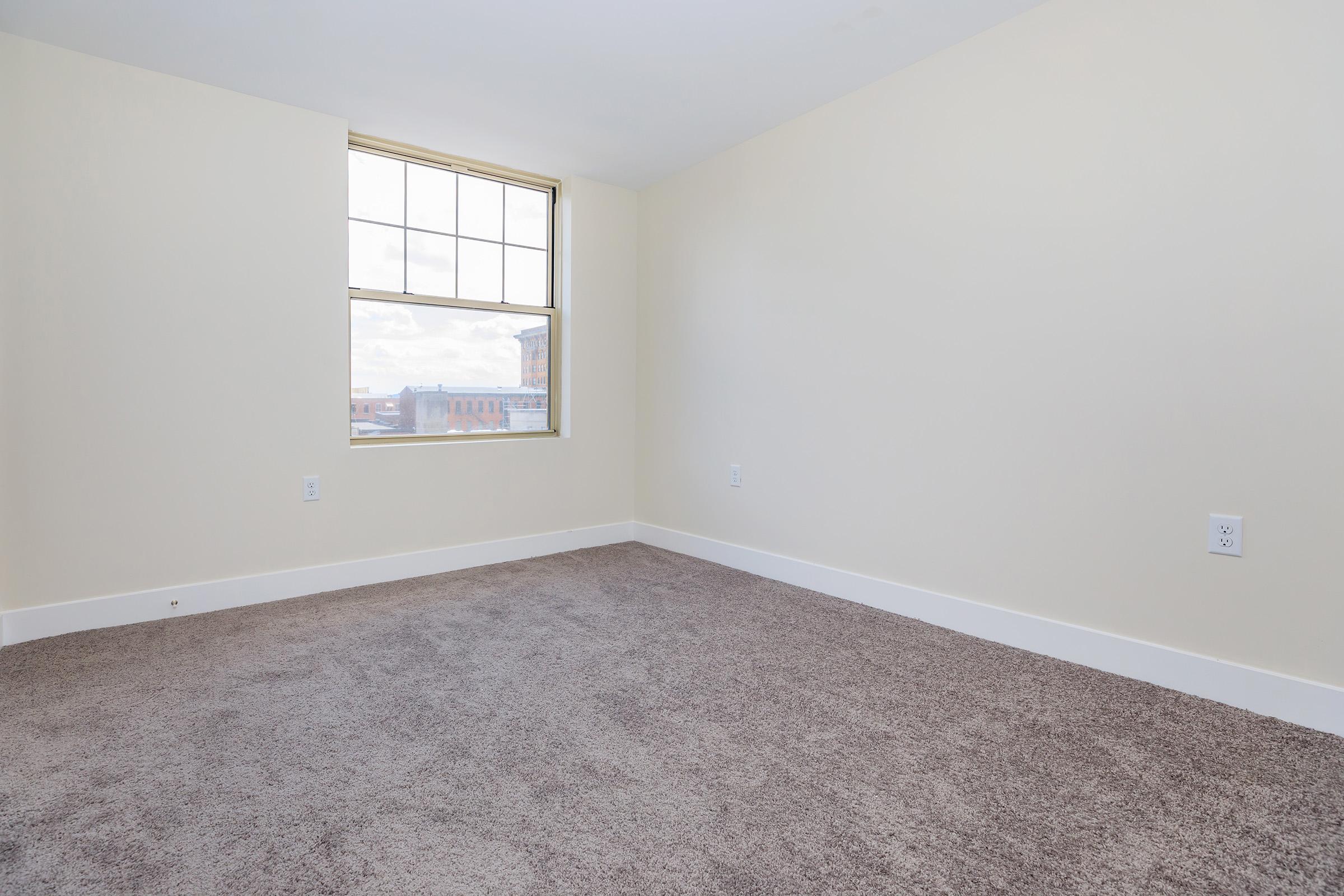 A clean, empty room featuring light-colored walls and a large window allowing natural light. The floor is covered with beige carpet, and there are no furniture or decorations present, creating a spacious and minimalist look. The window offers a view of a building exterior.