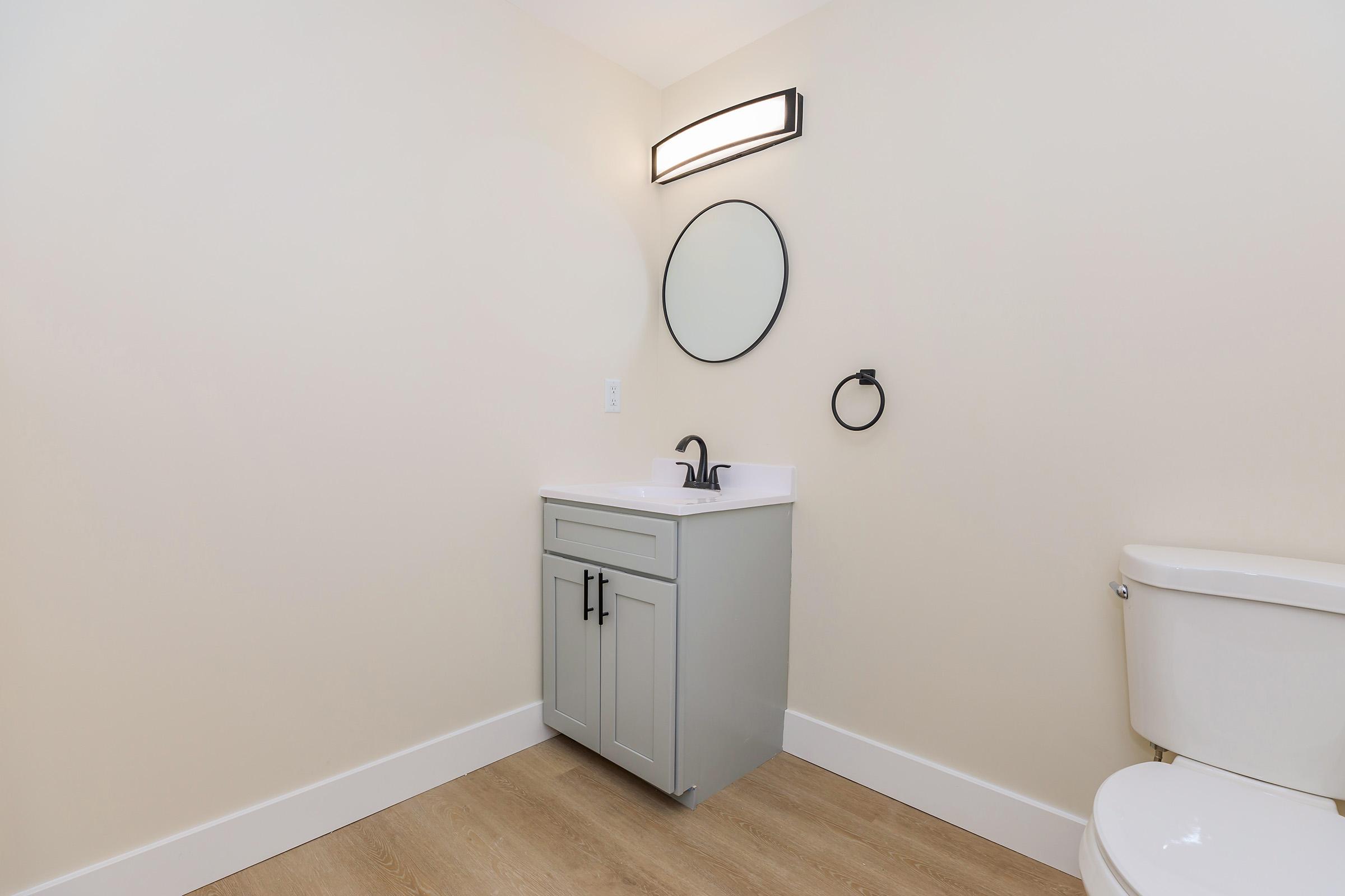 A modern bathroom featuring a light-colored wall and wood-like flooring. It includes a gray vanity with a sink, a round mirror above the sink, and a black faucet. To the right, there is a white toilet, and a black towel ring is mounted on the wall next to the vanity. The overall design is minimalistic and contemporary.