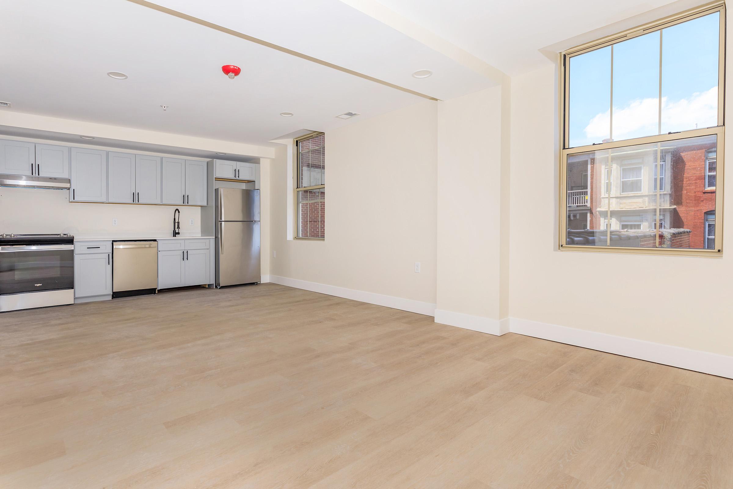 Spacious modern apartment interior featuring an open kitchen with stainless steel appliances, light-colored cabinetry, and a large living area with hardwood flooring. Large windows provide natural light, complementing the neutral wall colors.