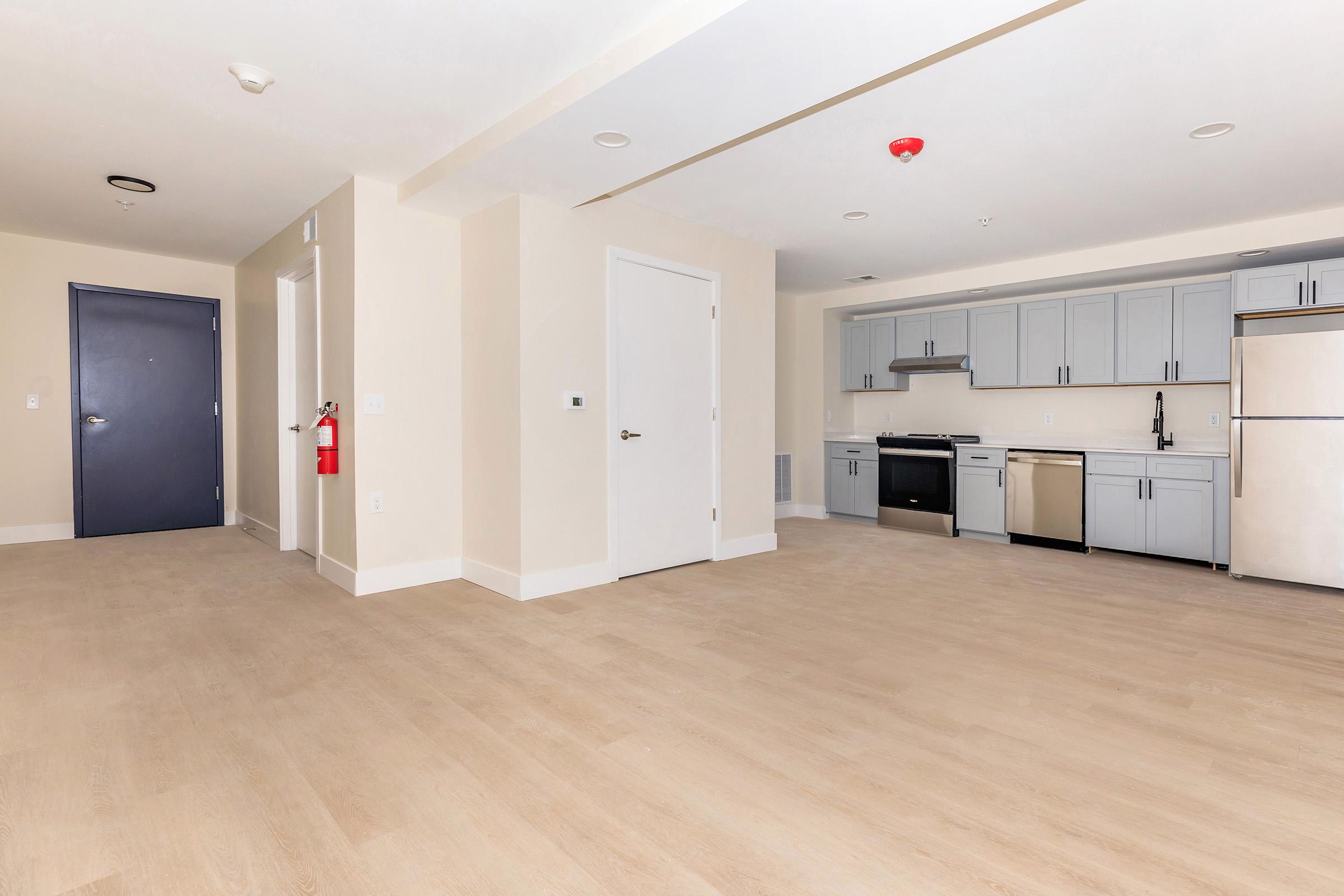 Spacious modern kitchen with stainless steel appliances, including a refrigerator, stove, and dishwasher. Light-colored wood flooring throughout. The room features neutral walls and ample natural light, with a door leading to another area and a fire extinguisher mounted on the wall.