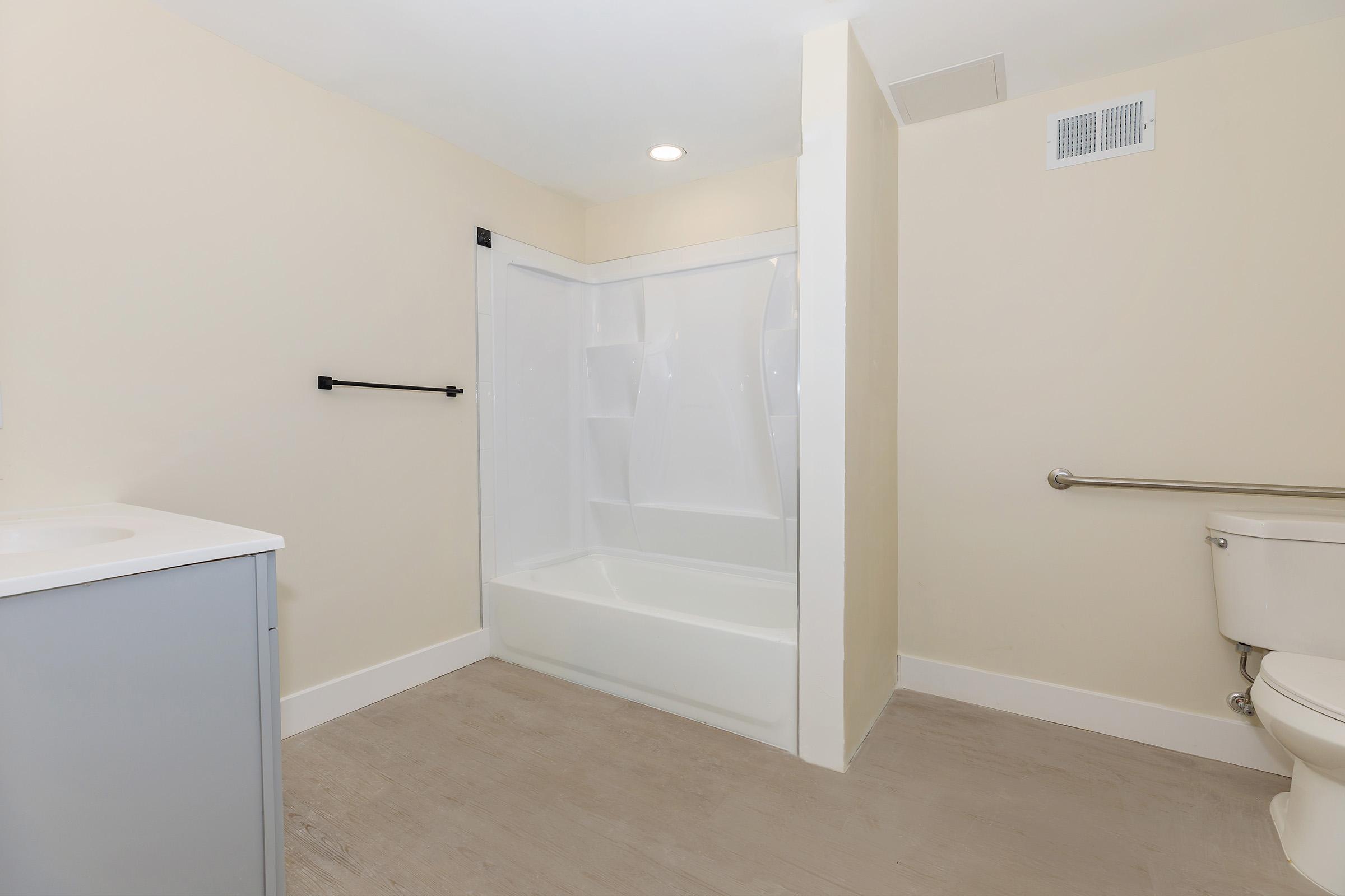 A clean, minimalistic bathroom featuring a bathtub with a shower, a toilet, and a small vanity. The walls are painted a light color, and there is a grab bar next to the toilet for accessibility. The floor has a simple, neutral finish.