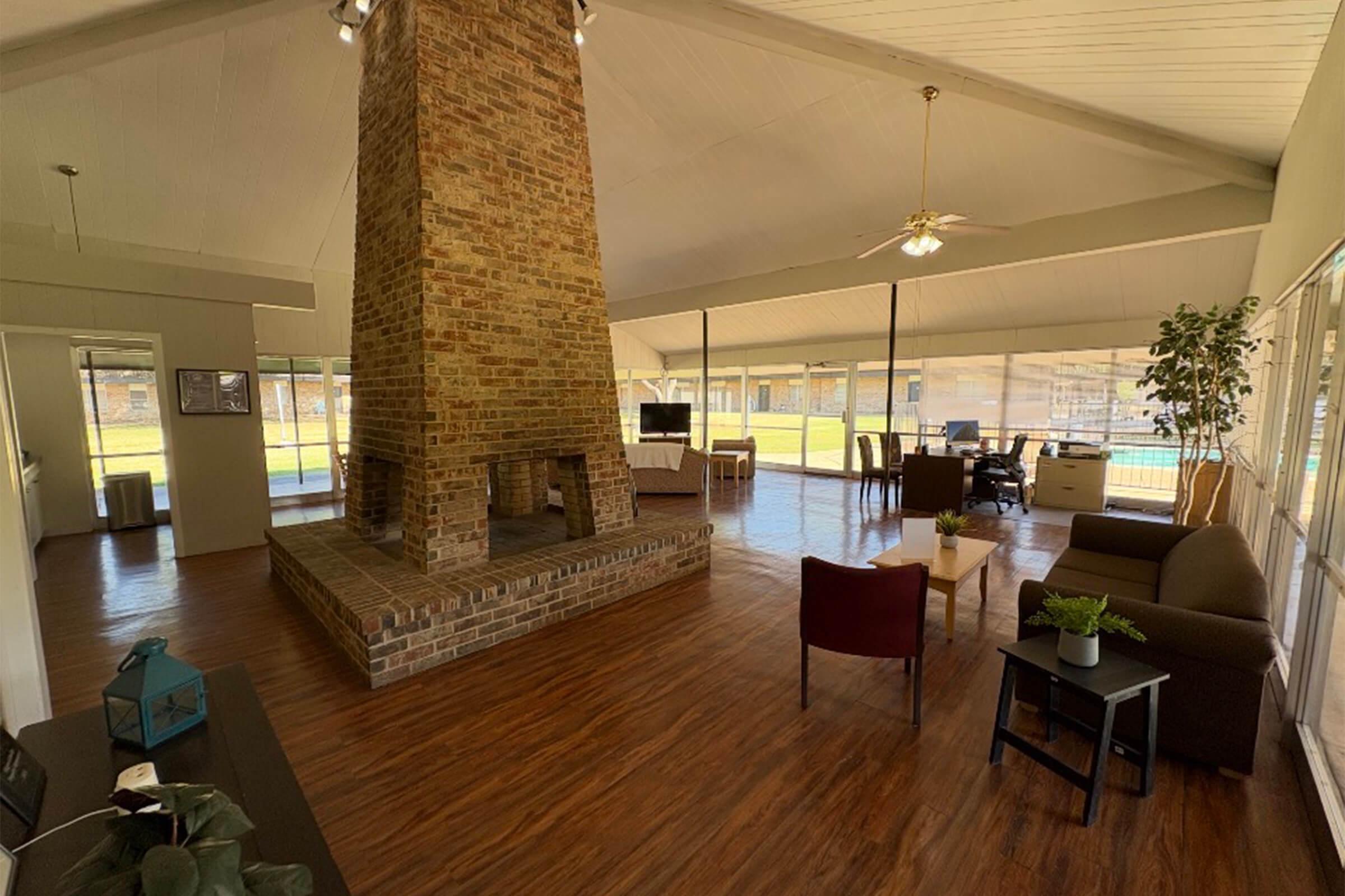 Spacious interior of a modern living area featuring a large brick fireplace as a focal point. The room has wooden flooring, a high ceiling, and large windows allowing natural light. Furnishings include a brown armchair, a small table, and a green plant. A view of an outdoor pool is visible through the windows.