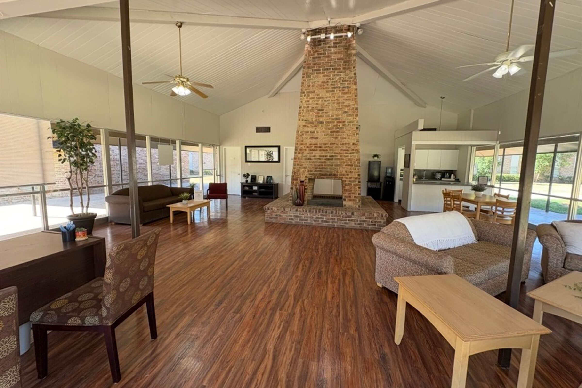 A spacious, modern interior of a living area featuring hardwood floors, large windows, and a high ceiling. A prominent brick fireplace is at the center, surrounded by comfortable seating, including sofas and chairs. A dining area is visible in the background, along with a kitchenette.